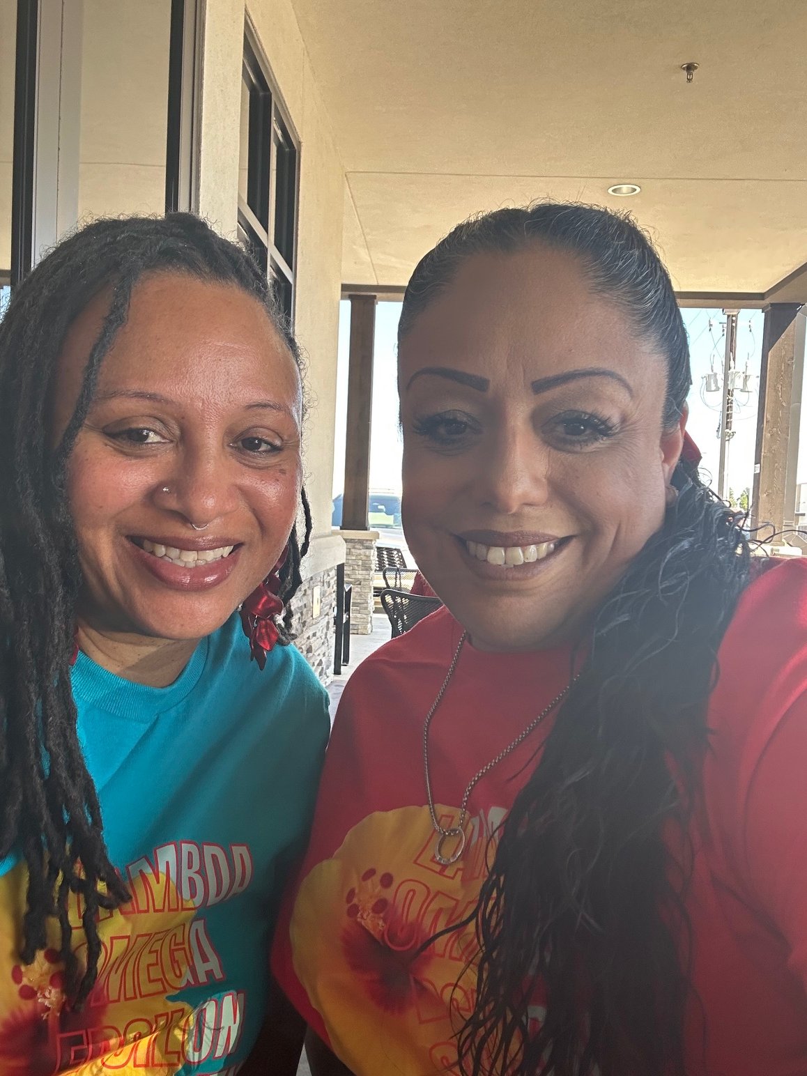 Two women smiling for a selfie at an outdoor cafe, with a beige wall and large windows behind them.