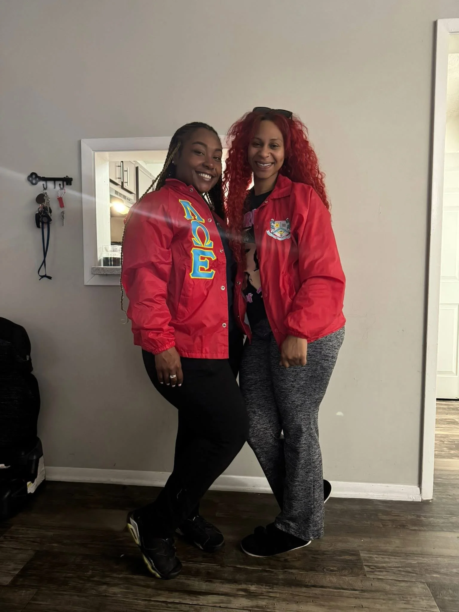 Two women smiling and standing close together indoors. One woman is wearing a red jacket with Greek letters and the other is wearing a red jacket with a crest. They are standing in front of a plain wall with a small mirror and some keys hanging on a 