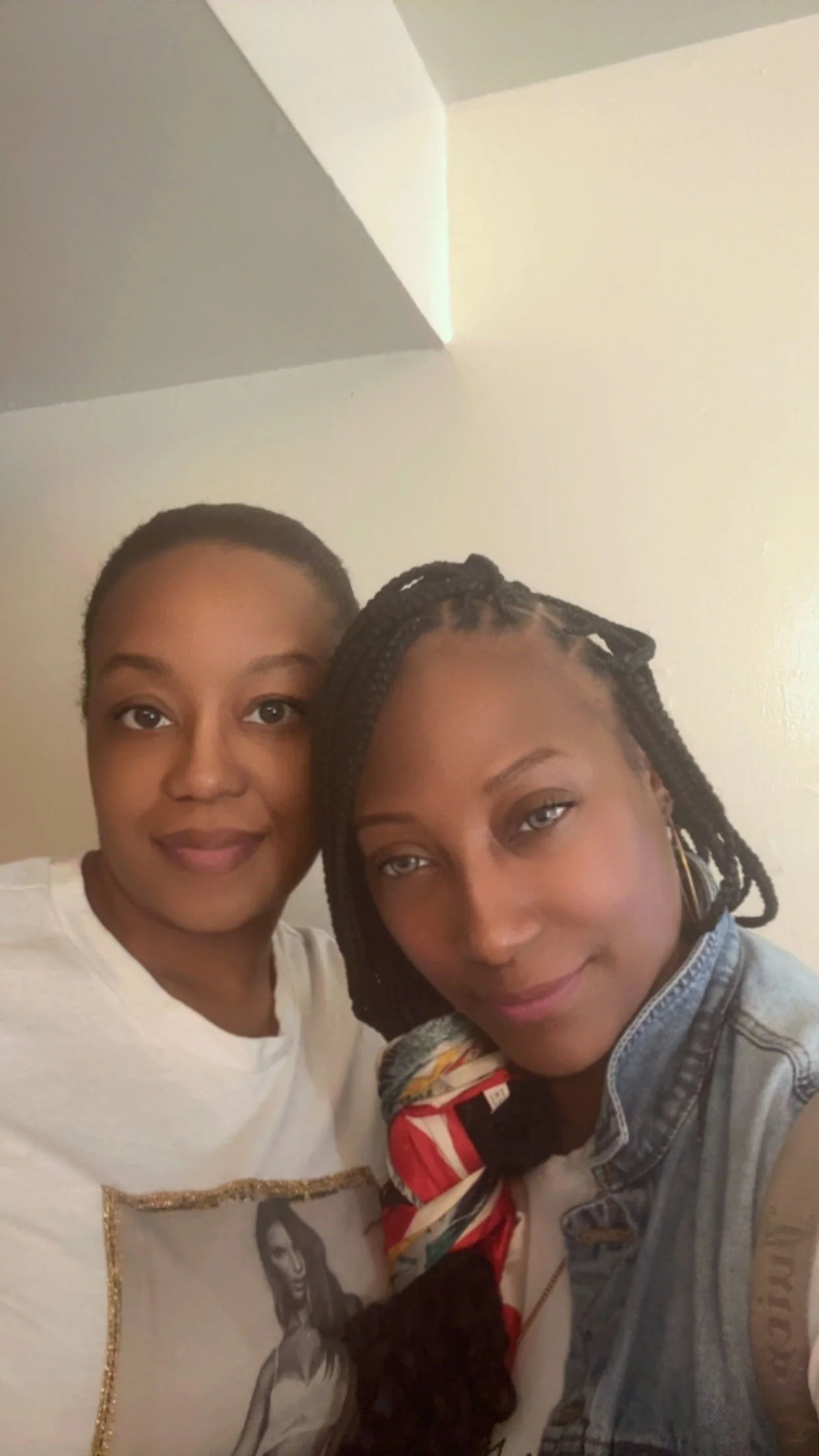 Two women taking a selfie indoors against a beige wall, one with short natural hair and the other with long braided hair wearing a denim jacket and colorful scarf.