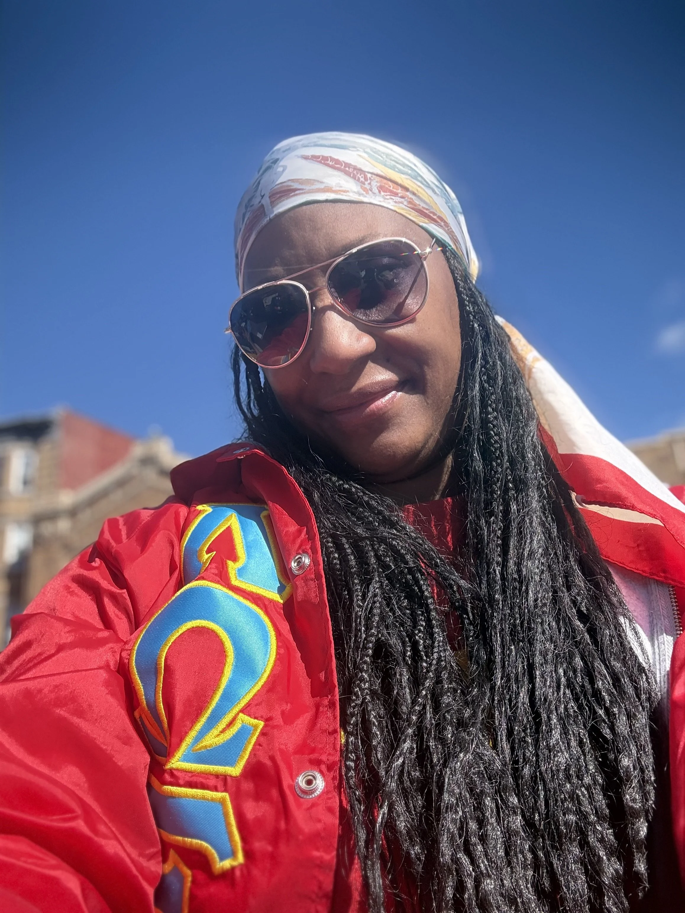 Close-up of a woman wearing sunglasses, a colorful headscarf, a red jacket with a bright patch, and long black curly hair, against a clear blue sky and some rooftops in the background.