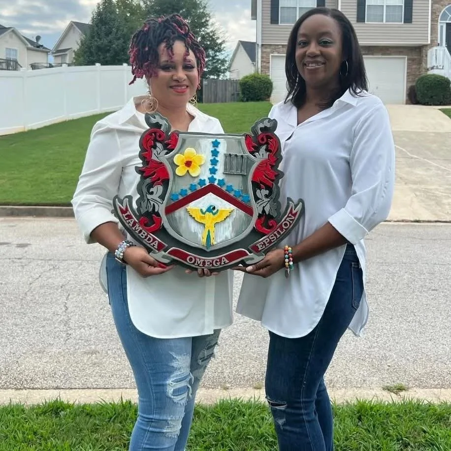 Two women standing outside on a sidewalk, holding a shield-shaped fraternity crest with the words 'Zeta Phi Beta' and 'Omega Psi Phi'. Both women are wearing white shirts and jeans, and are smiling at the camera. Background includes a grassy yard, a white fence, and houses.
