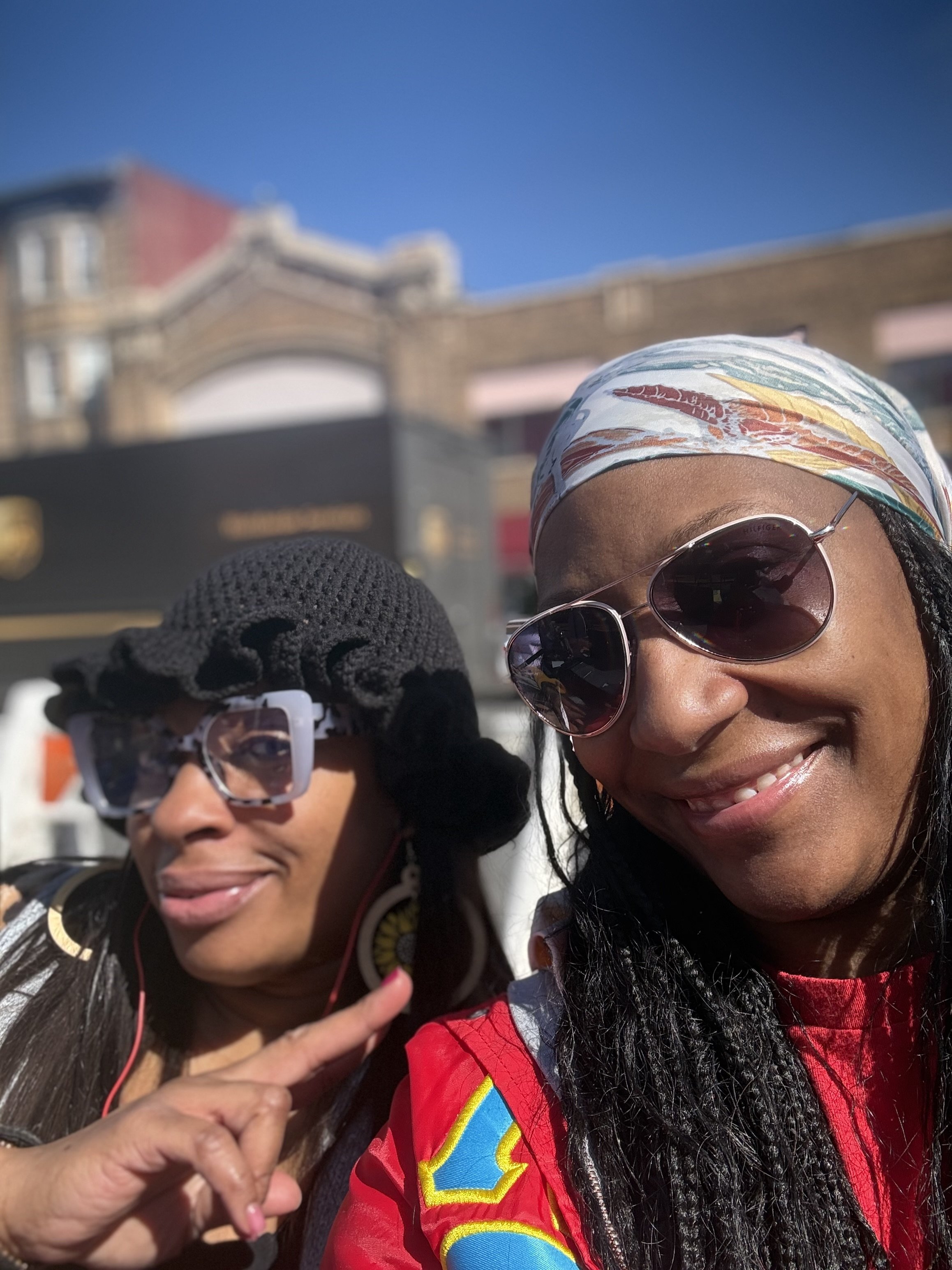 Two women smiling outdoors on a sunny day with a historic building in the background. One woman wears sunglasses and a colorful headscarf, the other wears glasses and has long dark hair.