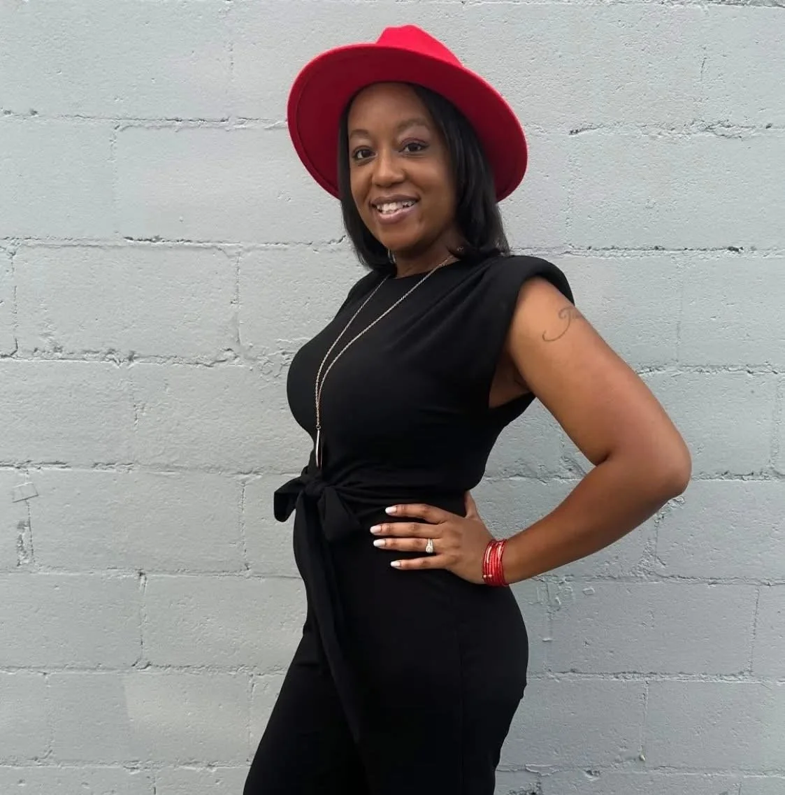 Woman wearing a red hat and black outfit, standing against a gray brick wall, smiling with her hand on her hip.