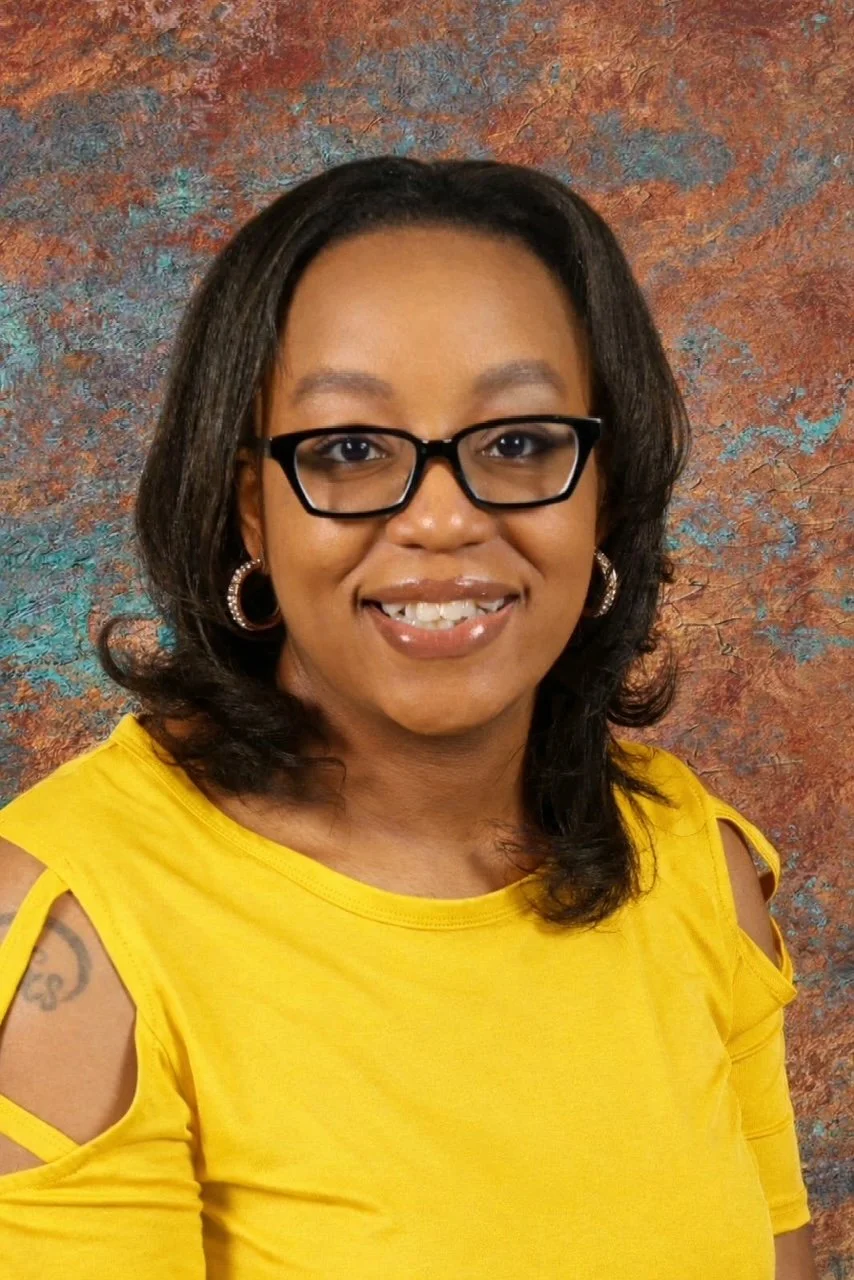 A woman with glasses and hoop earrings wearing a yellow top, standing in front of a textured, multicolored background.