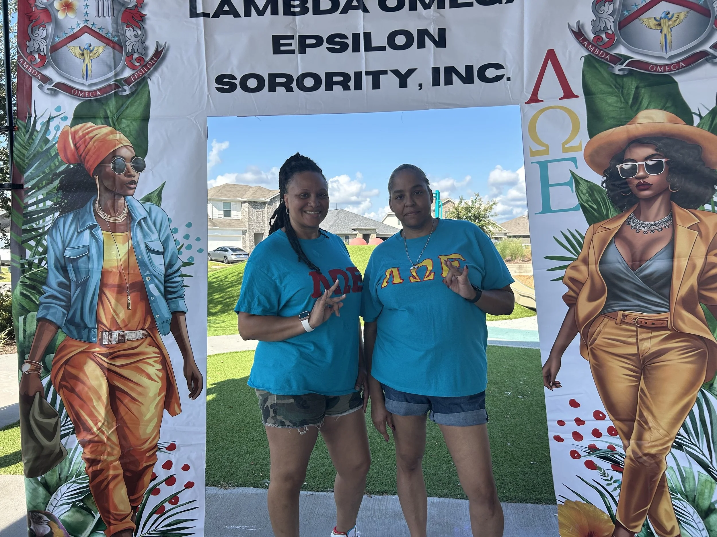 Two women standing underneath a banner that reads 'Lambda Omega Epsilon Sorority, Inc.' with colorful illustrations of women and tropical leaves.