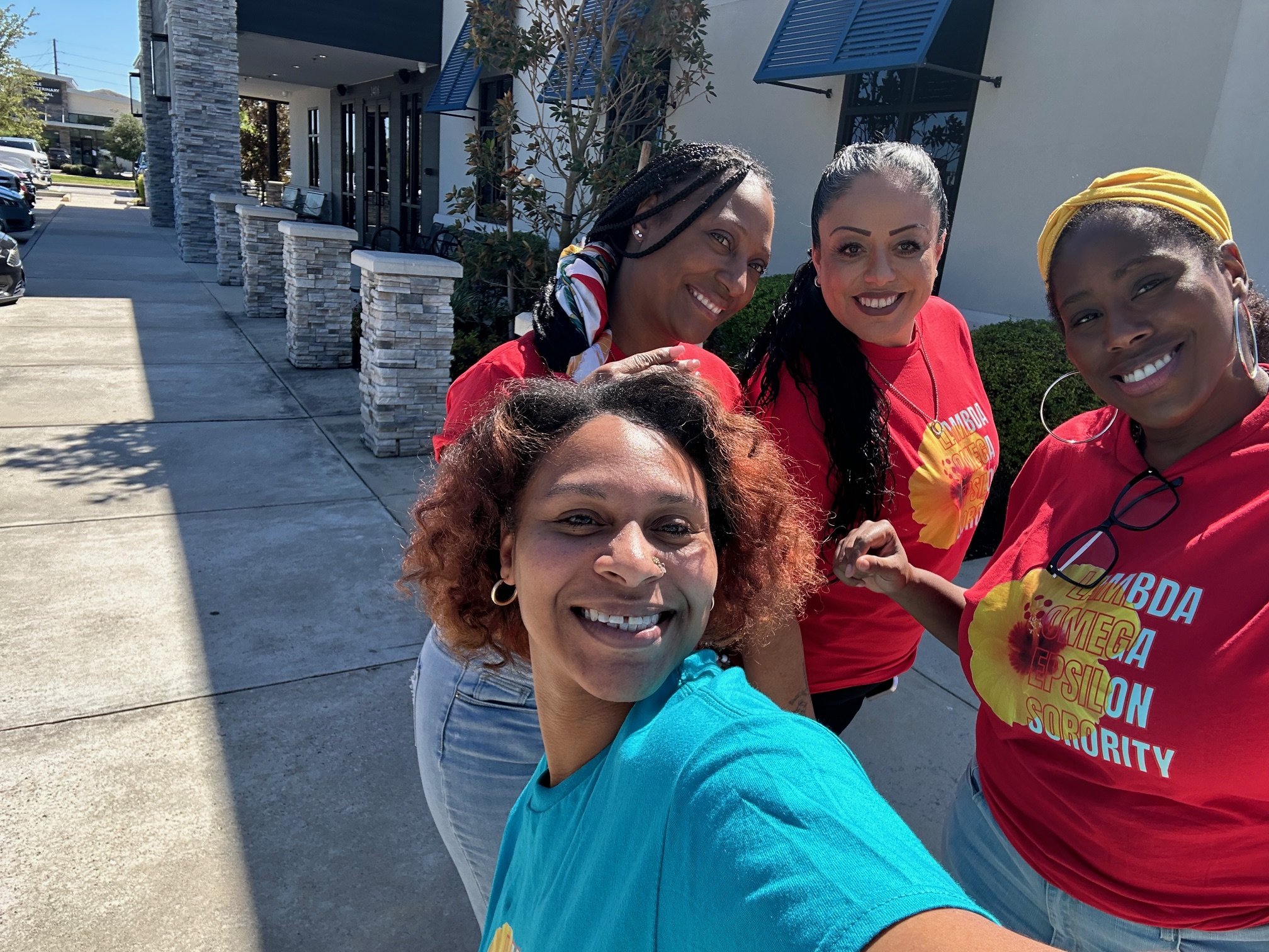 Group of women smiling outdoors in front of a building, wearing red T-shirts with colorful text, during daytime.