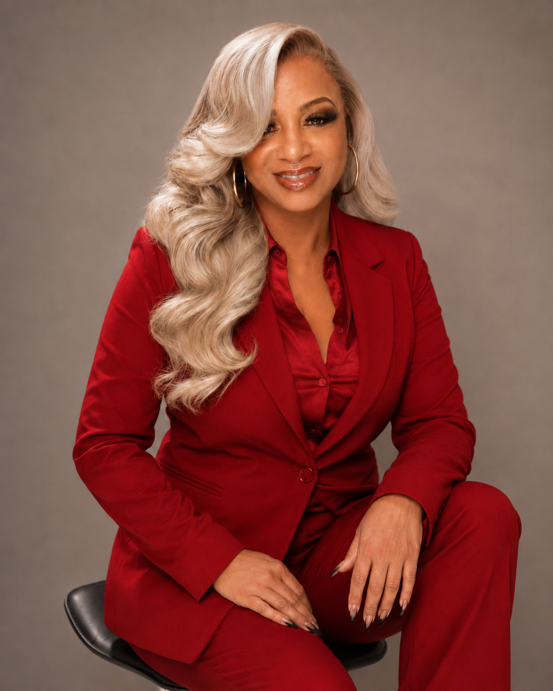Woman with long, blonde, wavy hair wearing a red business suit and sitting on a stool, smiling at the camera.