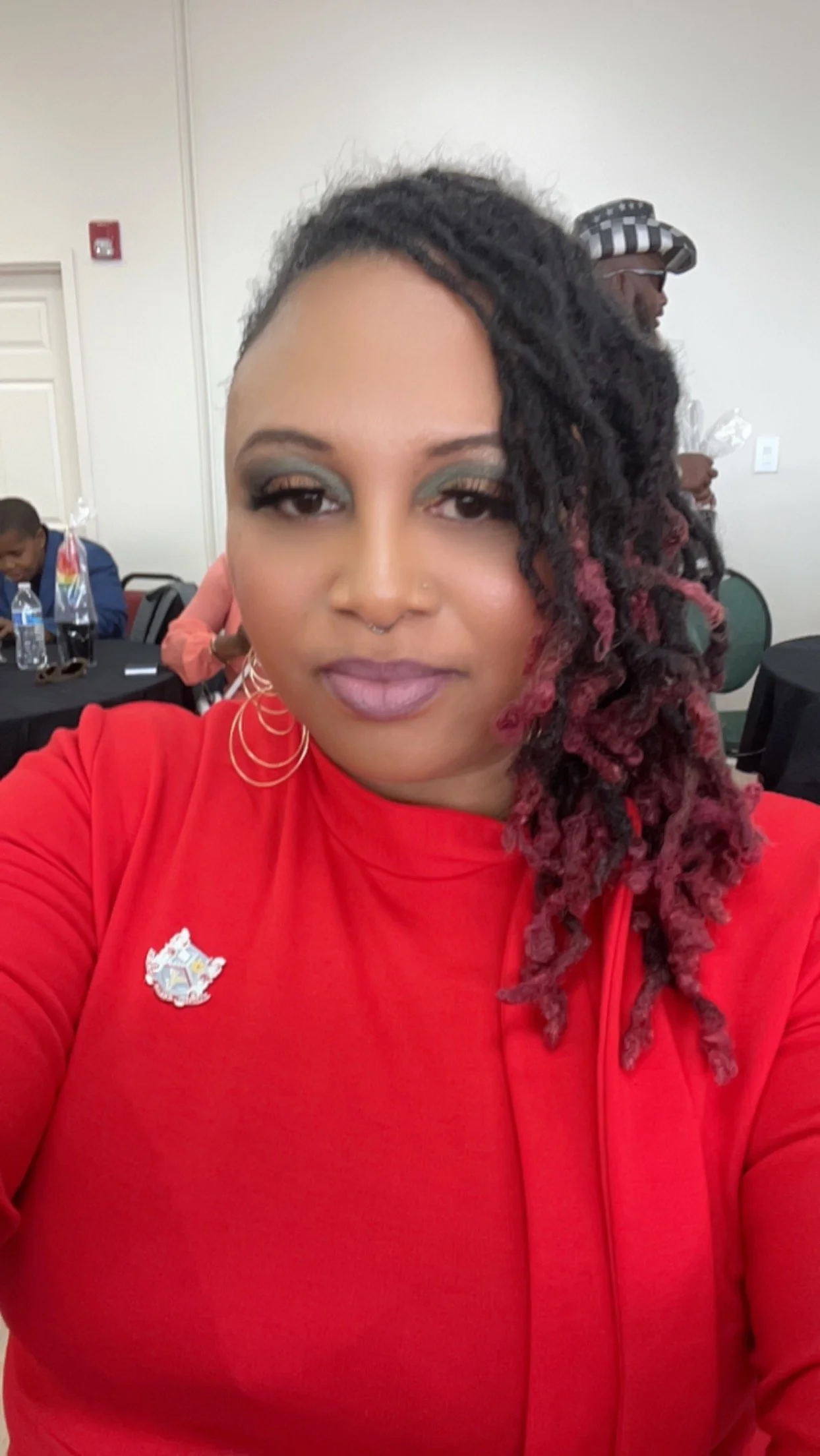 A woman with shoulder-length dreadlocks, some dyed pink, is taking a selfie at an indoor event. She is wearing a red shirt with a small embroidered emblem and large hoop earrings. In the background, there are people sitting at tables, some with water