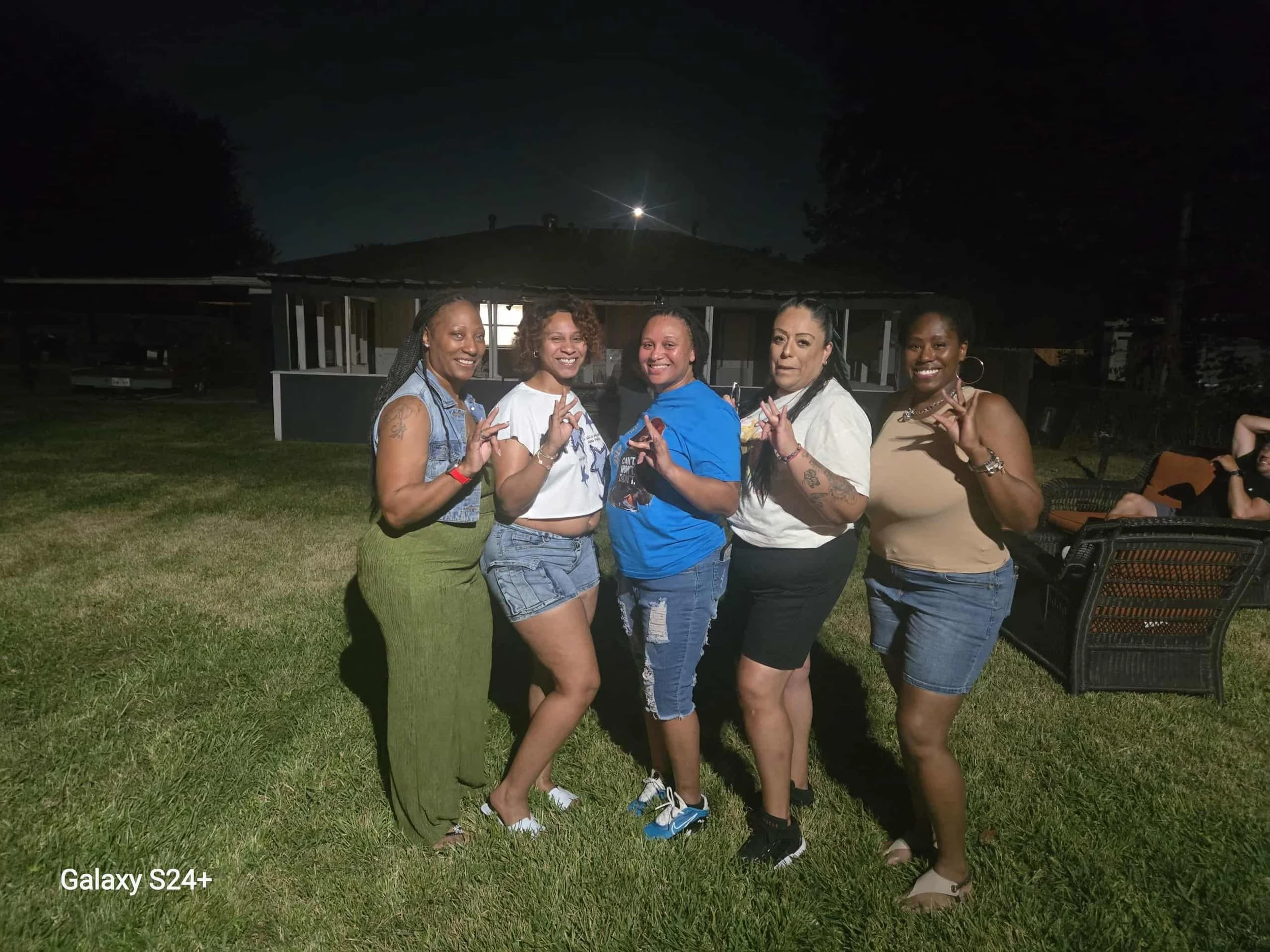 Five women standing closely together outside at night, smiling, making peace signs; house and moonlit sky in background.