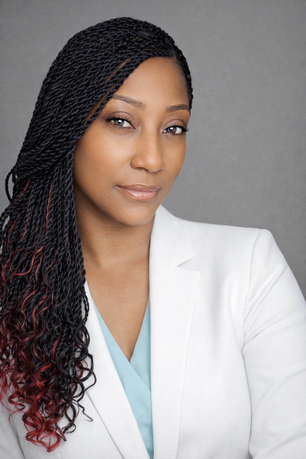 A woman with long braided hair, wearing a white blazer and a light blue top, looking at the camera with a neutral expression, against a gray background.