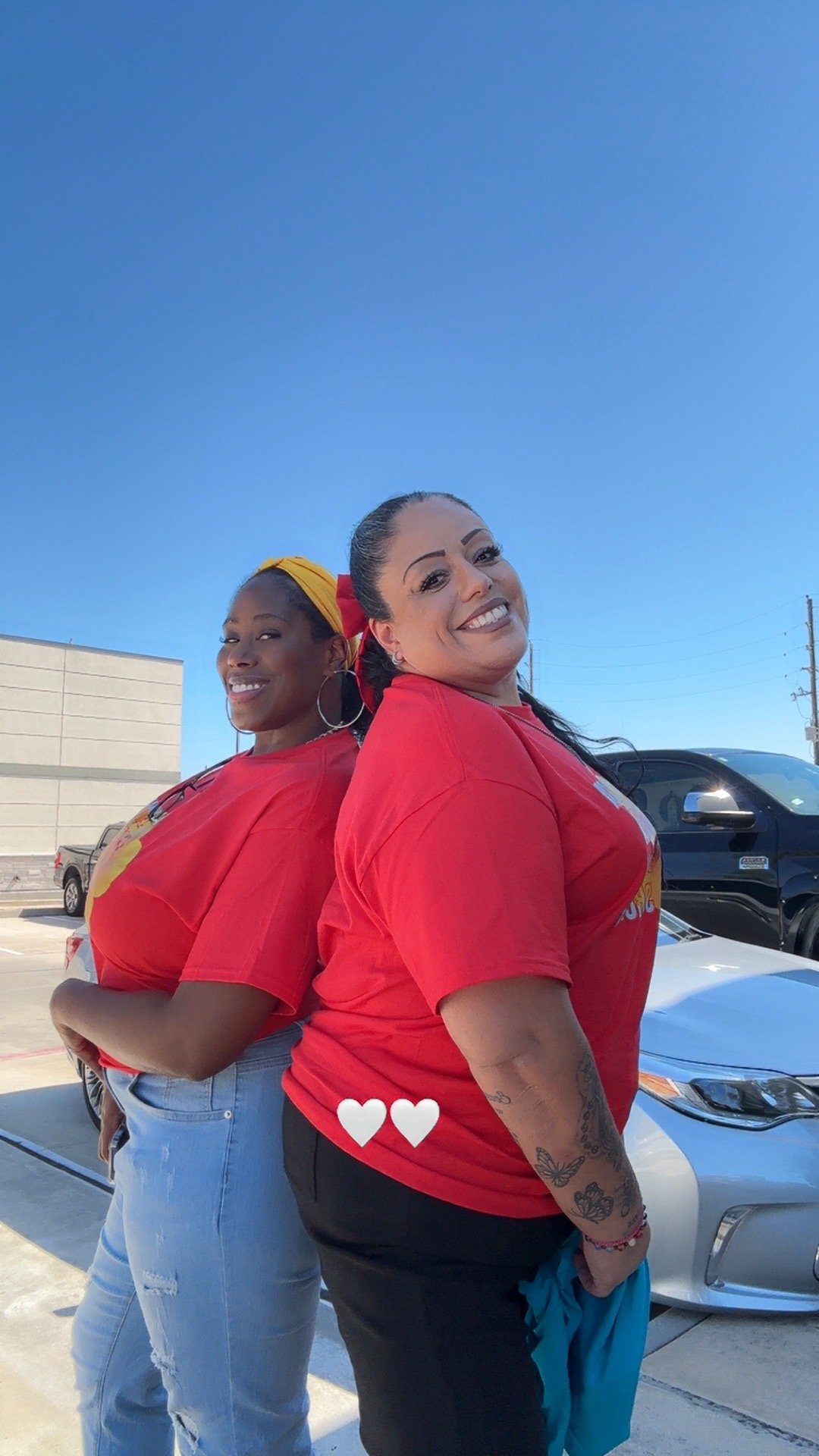 Two women standing back to back outdoors, smiling, wearing red t-shirts, with parked cars in the background and a clear blue sky.