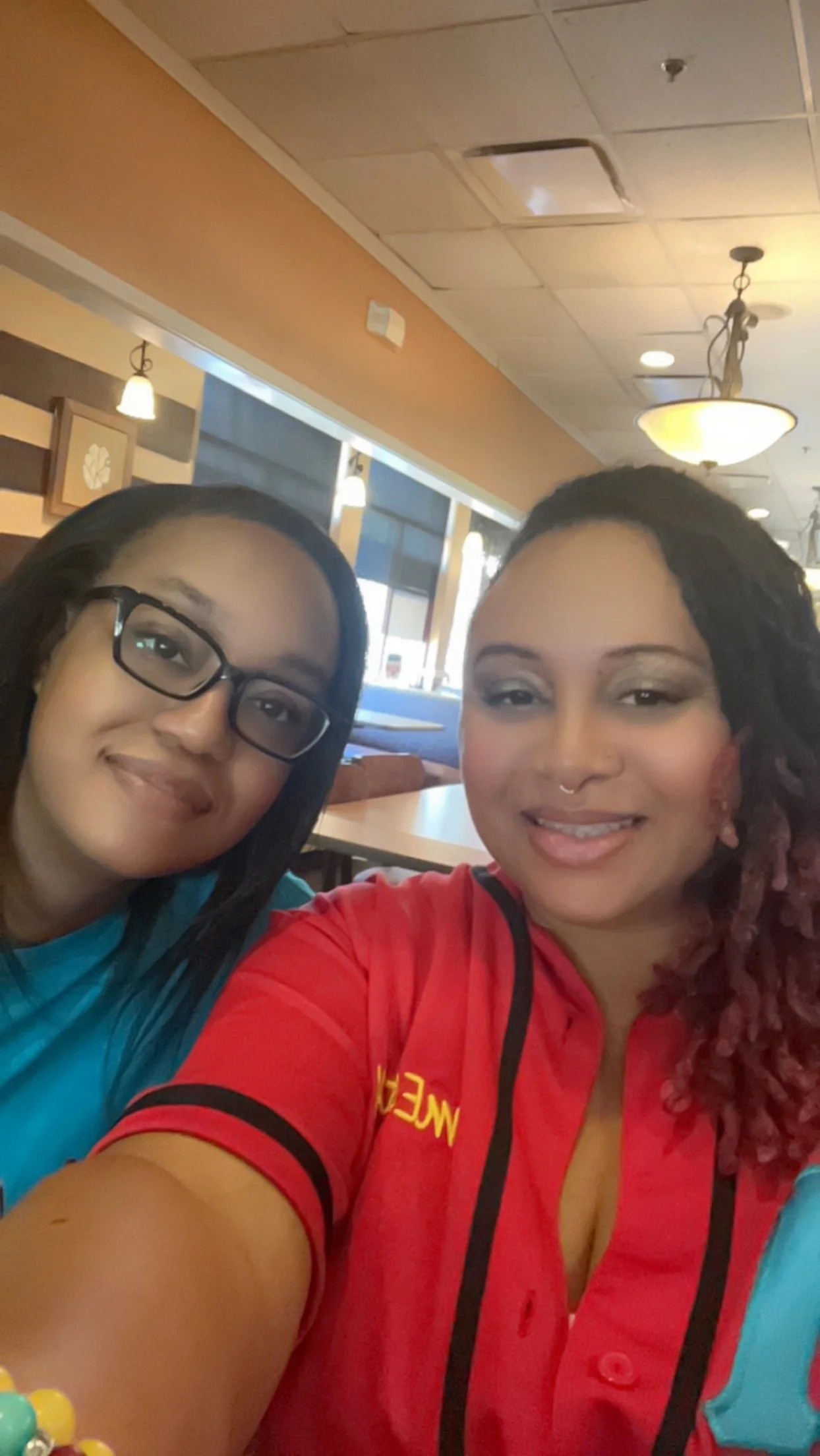 Two women taking a selfie inside a restaurant, smiling and sitting at a table with light-colored walls, ceiling lights, and decorative wall panels in the background.