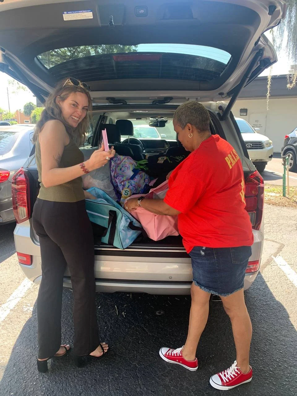 Two women are packing items into the back of a silver SUV parked in a parking lot. The woman on the left has long hair, is wearing a black sleeveless top and black pants, and is smiling while holding a pink item. The woman on the right has short hair, is wearing a red shirt, denim shorts, and red sneakers, and is placing items into the trunk.