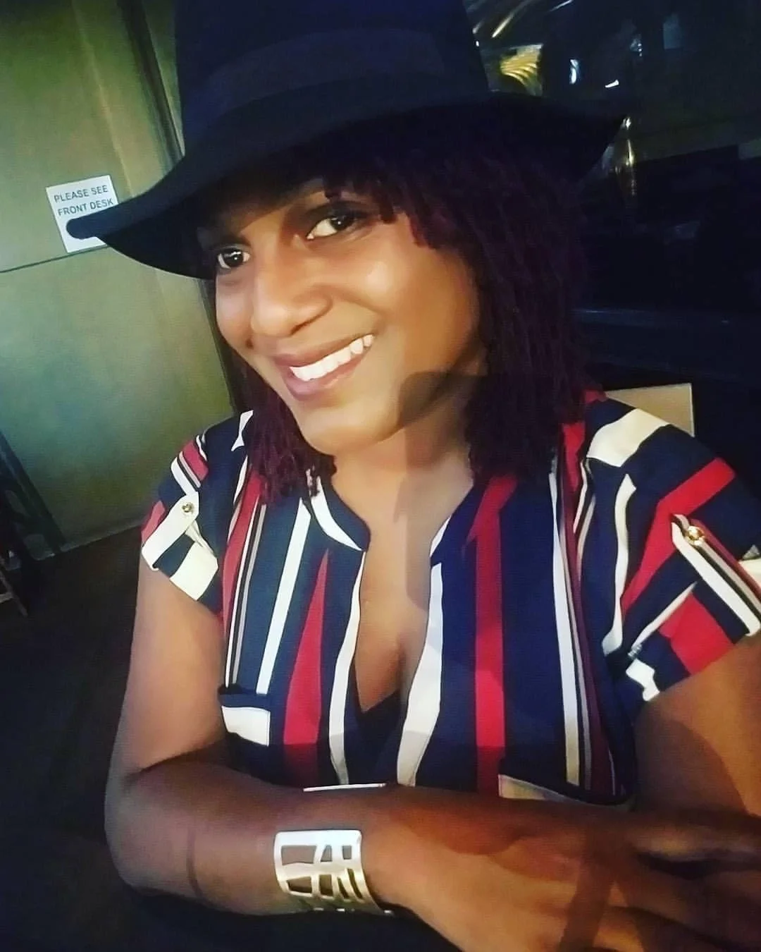 A woman with dark skin, shoulder-length curly hair, wearing a wide-brimmed black hat and a striped shirt with red, white, and blue colors, sitting at a table, smiling at the camera.