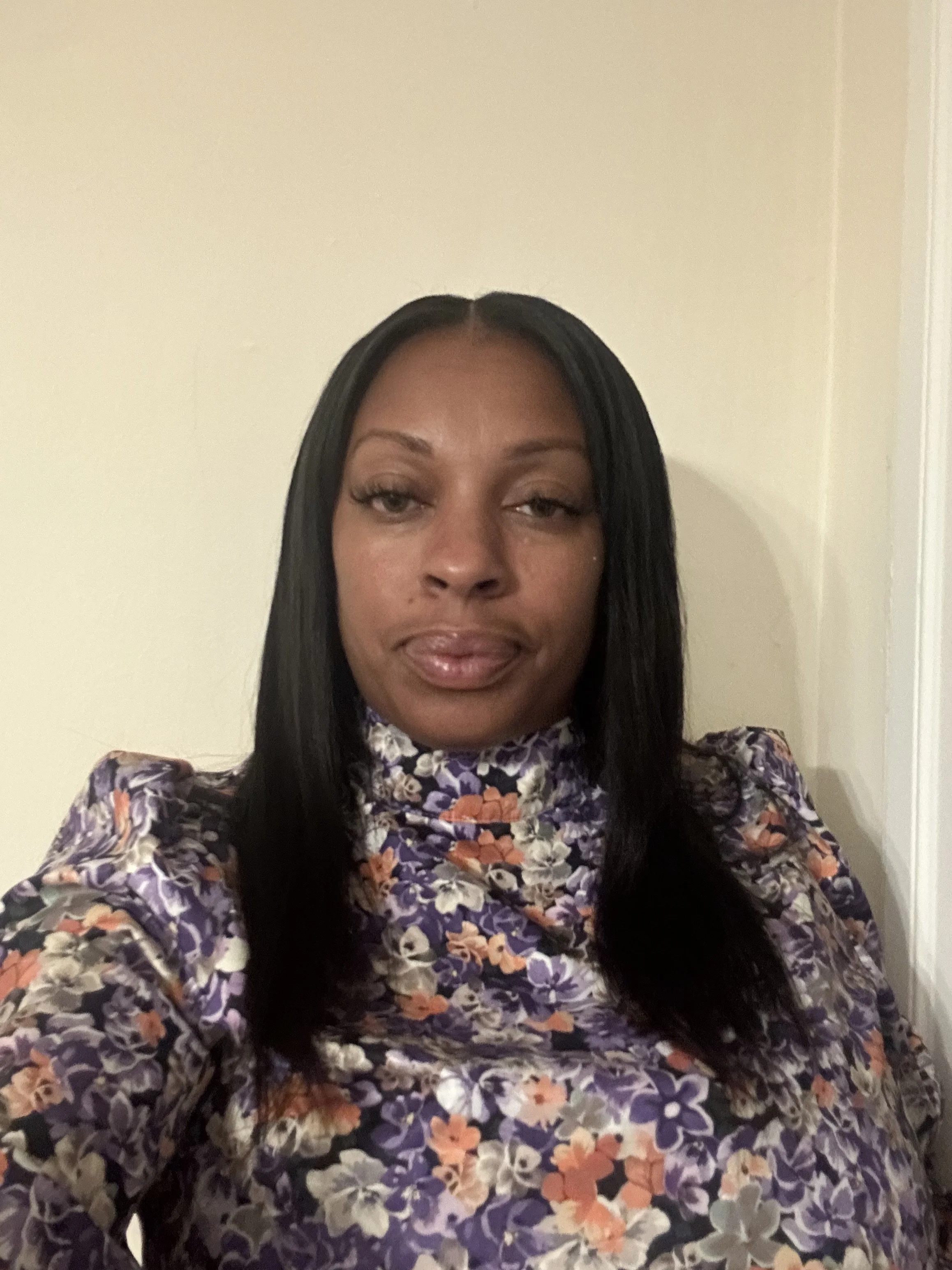 A woman with long black hair wearing a floral patterned top, taking a selfie inside a room with beige walls.