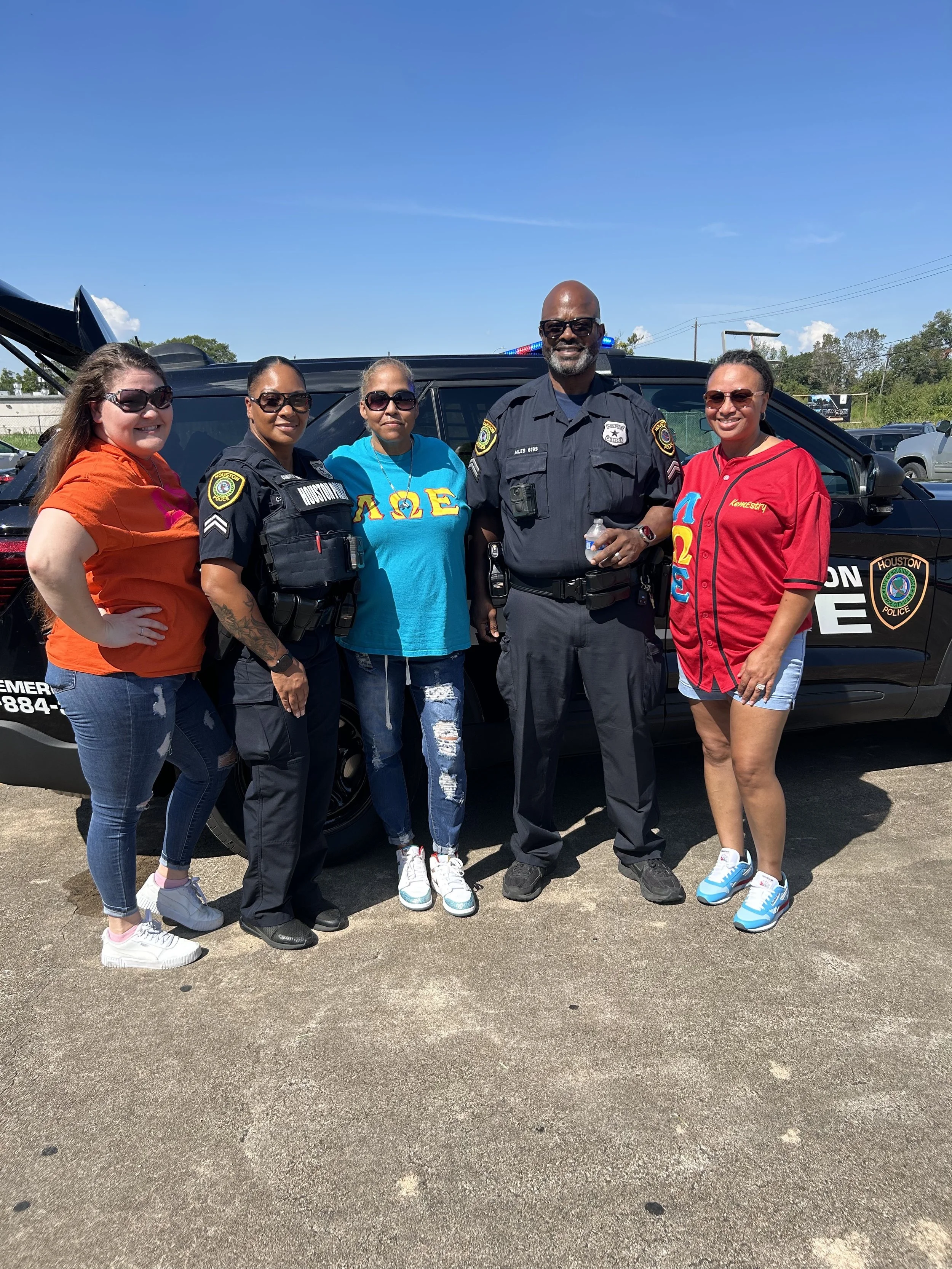 Group of five women and one police officer standing in front of a police vehicle outdoors on a sunny day.