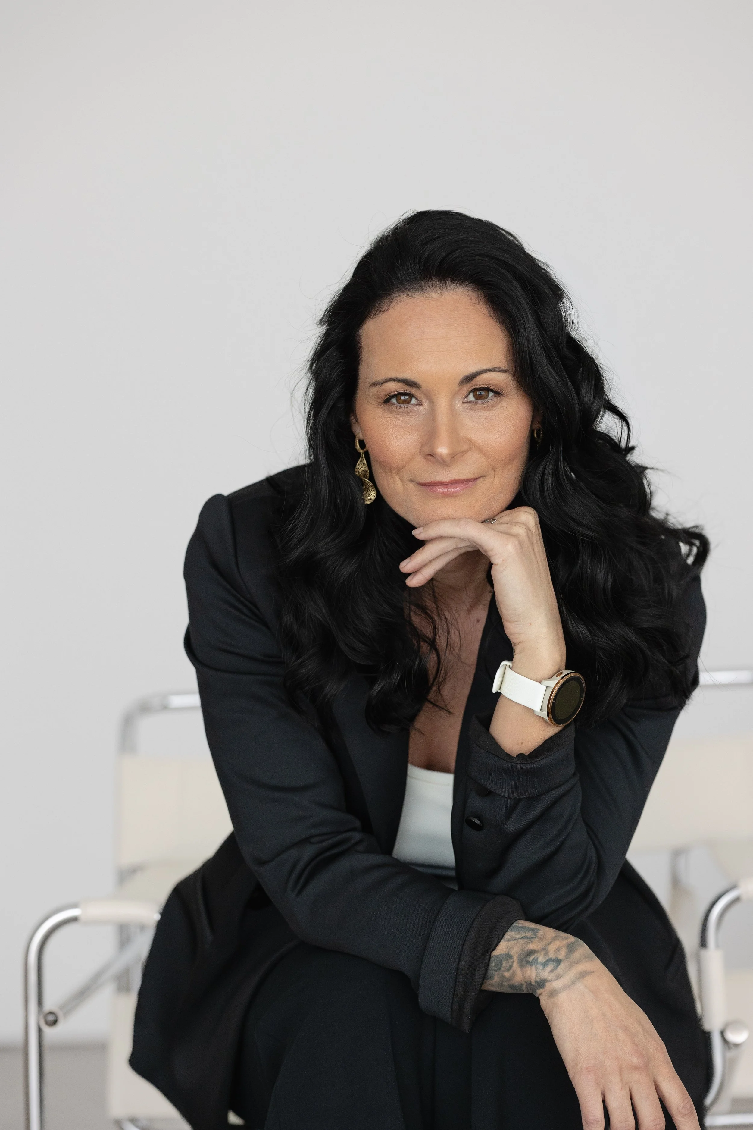 Femme avec cheveux noirs, portant un blazer noir, une montre blanche et une boucle d'oreille en or, assise sur une chaise en métal, posant avec le menton sur sa main, arrière-plan blanc.