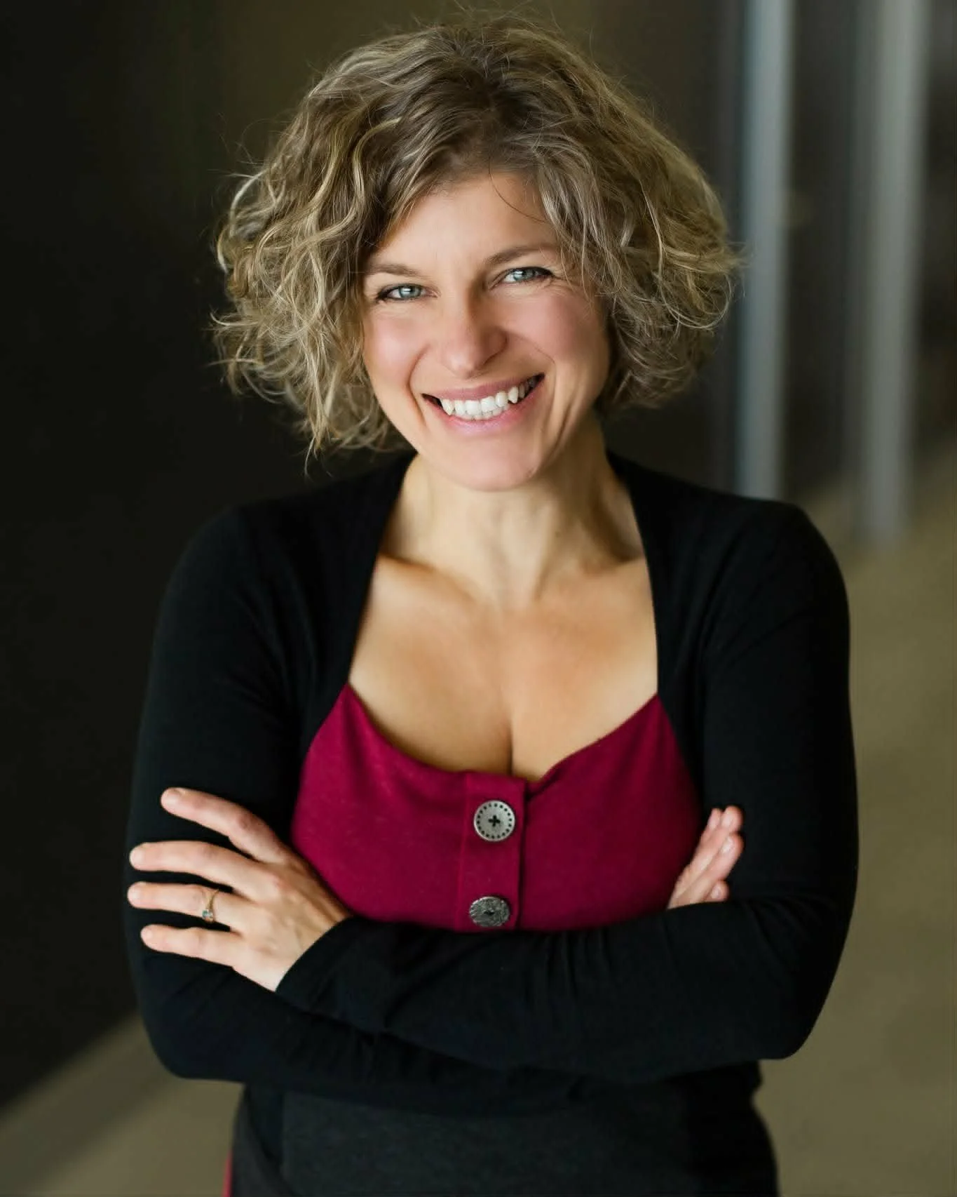 Femme souriante avec cheveux bouclés et vêtements colorés, bras croisés.