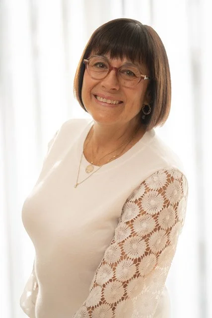 Une femme souriante avec des lunettes, portant une robe blanche avec des manches en dentelle, debout devant un rideau blanc.