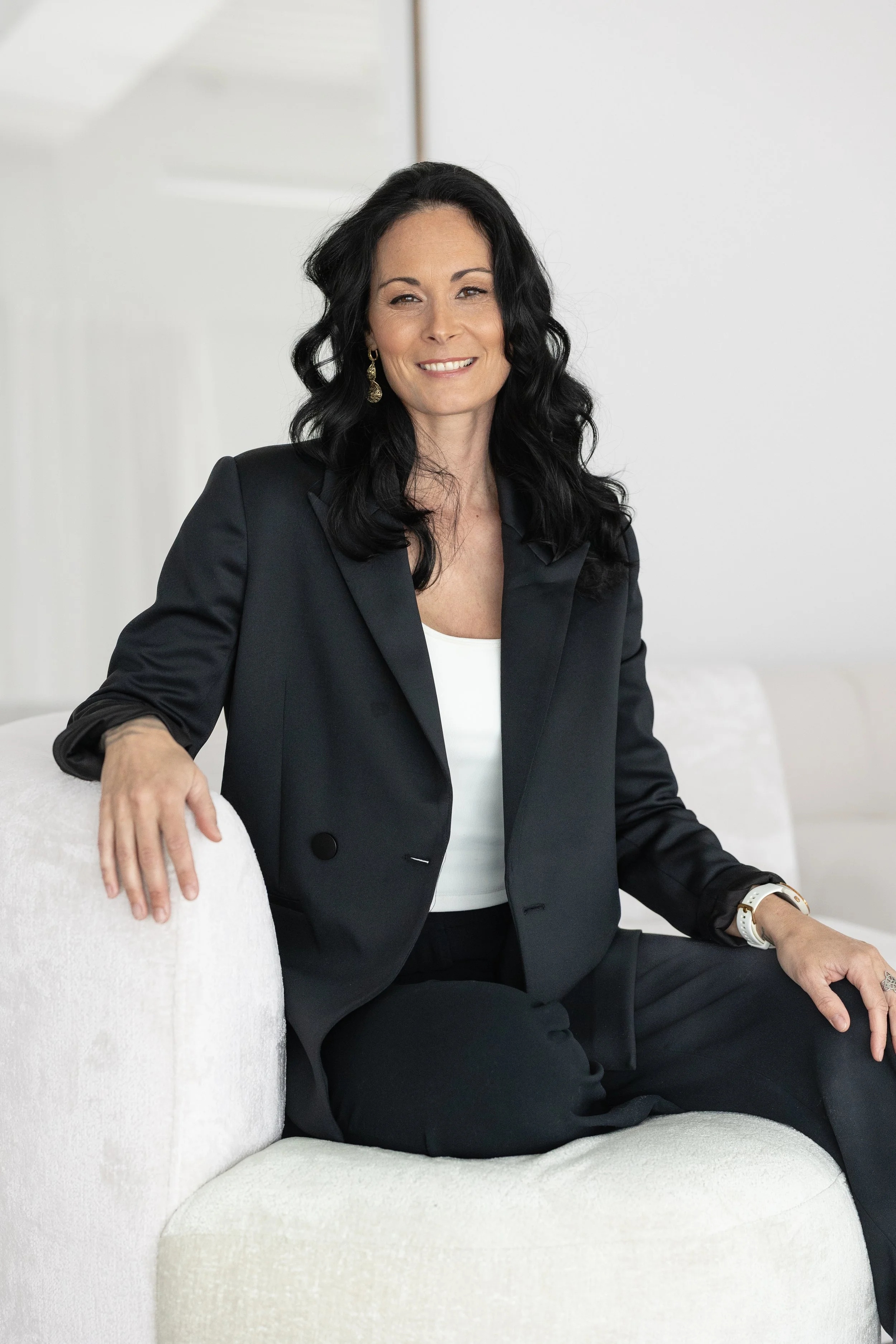 Femme assise sur un canapé blanc, portant un blazer noir, un haut blanc, et des boucles d'oreilles dorées, souriant vers l'objectif.