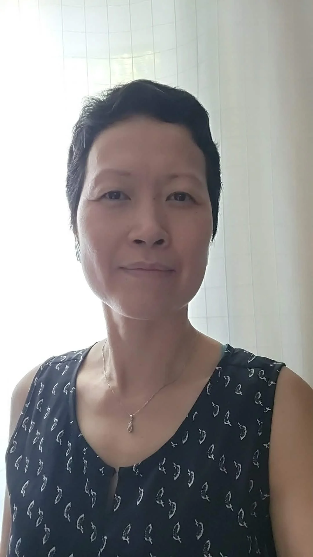 Une femme souriante portant une robe noire avec un motif blanc et une chaîne avec un pendentif, en intérieur avec un mur blanc en arrière-plan.