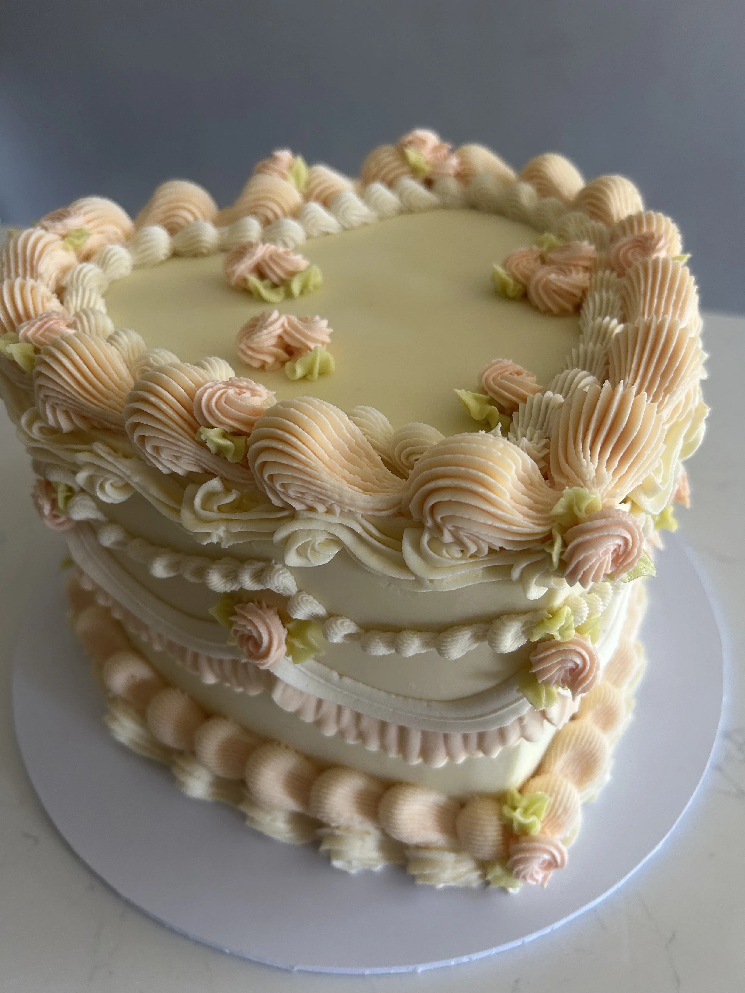 A heart-shaped cake decorated with pink, cream, and green swirled frosting on top and along the edges, placed on a white plate.