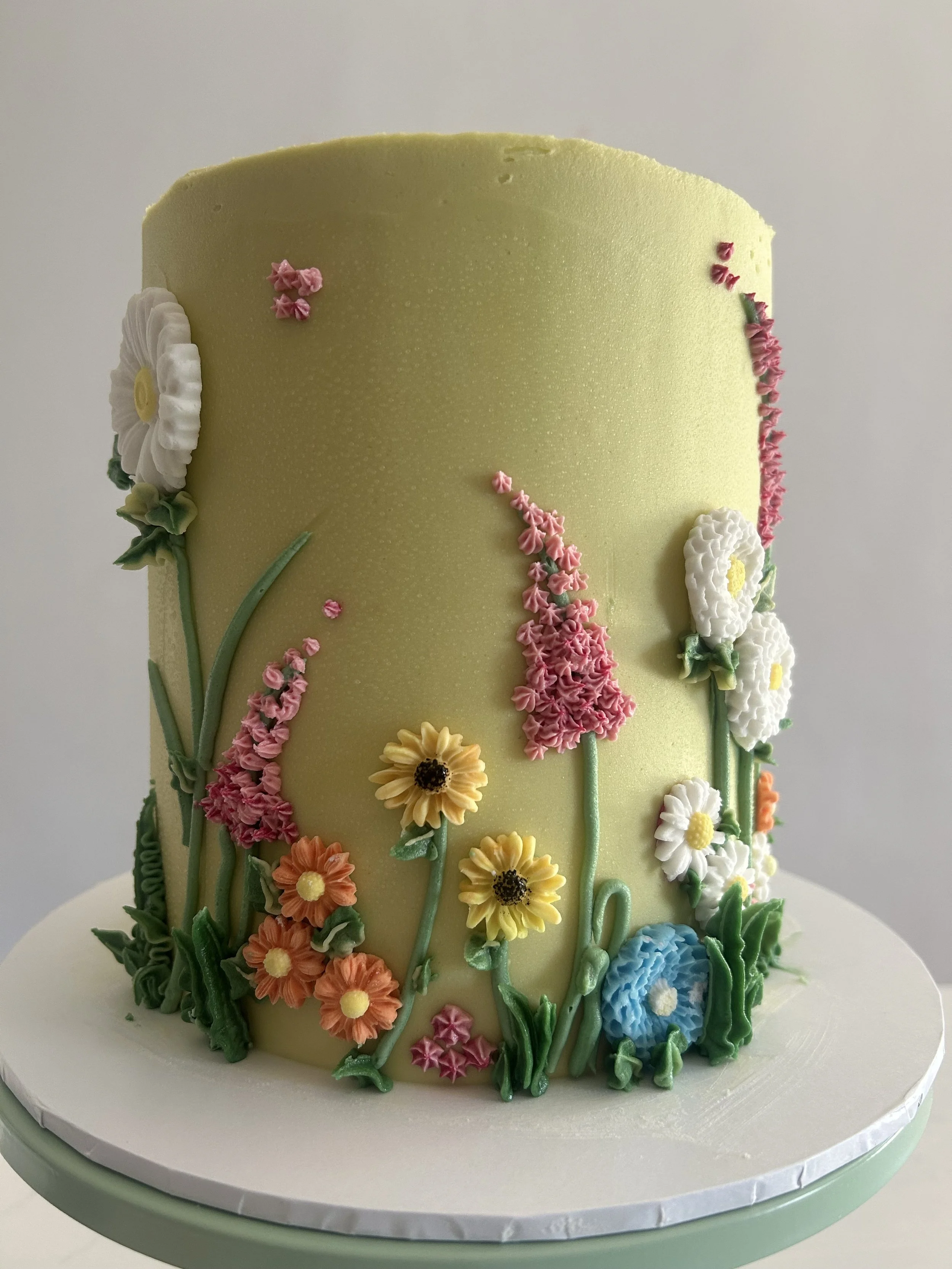 A cake decorated with intricate pastel-colored floral designs and green piping leaves on a white cake board.