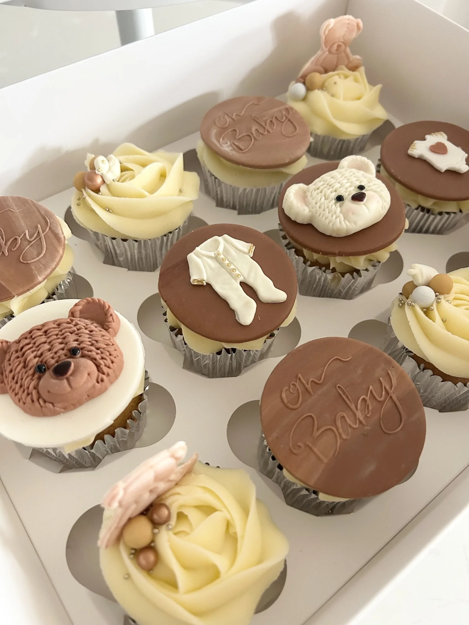 Box of decorative cupcakes with animal and baby-themed designs, including bears and baby clothes, with various icing decorations.