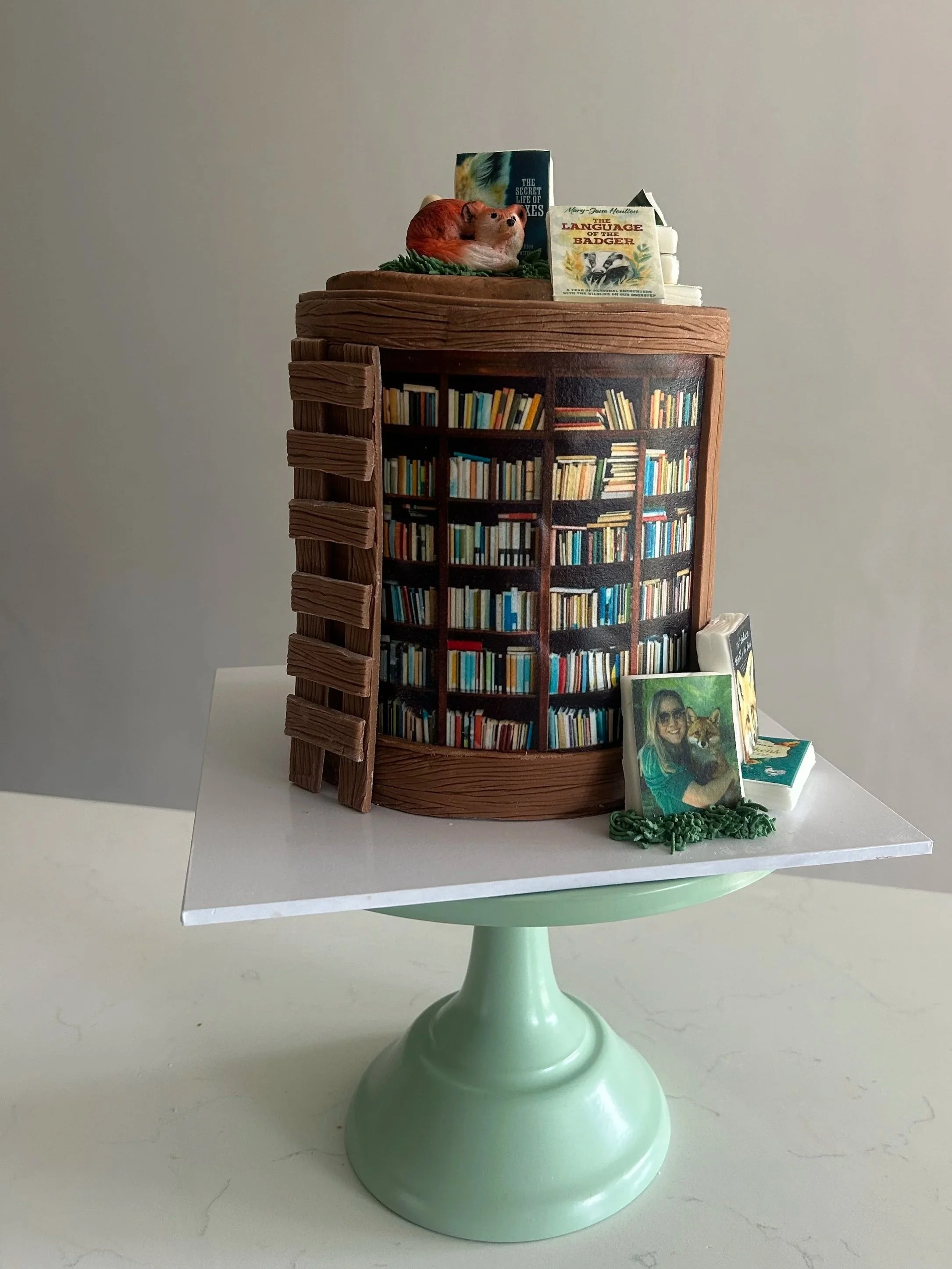 A table decorated with a bookshelf-themed cake, topped with animal figurines, books, and framed photos, placed on a white surface with a light-colored background.