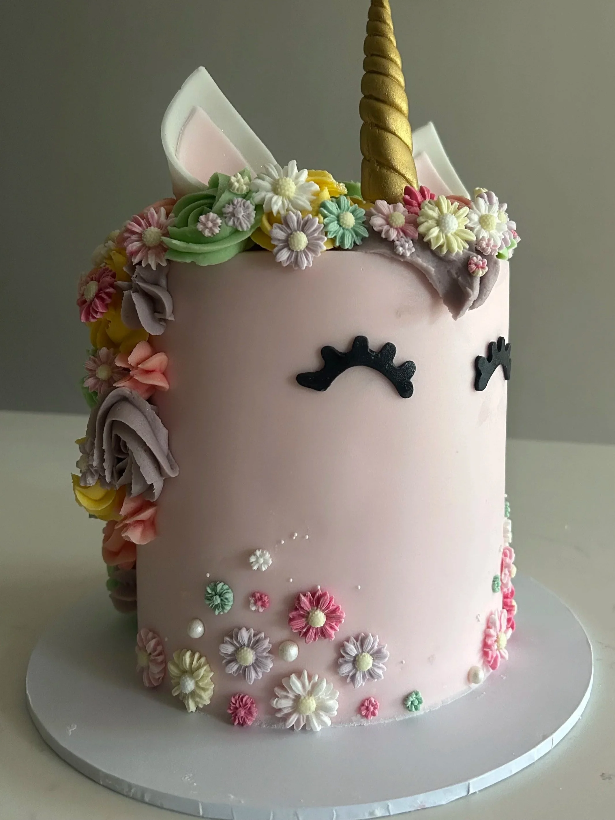 A pink unicorn-themed birthday cake decorated with colorful buttercream flowers, a gold unicorn horn, unicorn ears, and closed eyes with eyelashes.