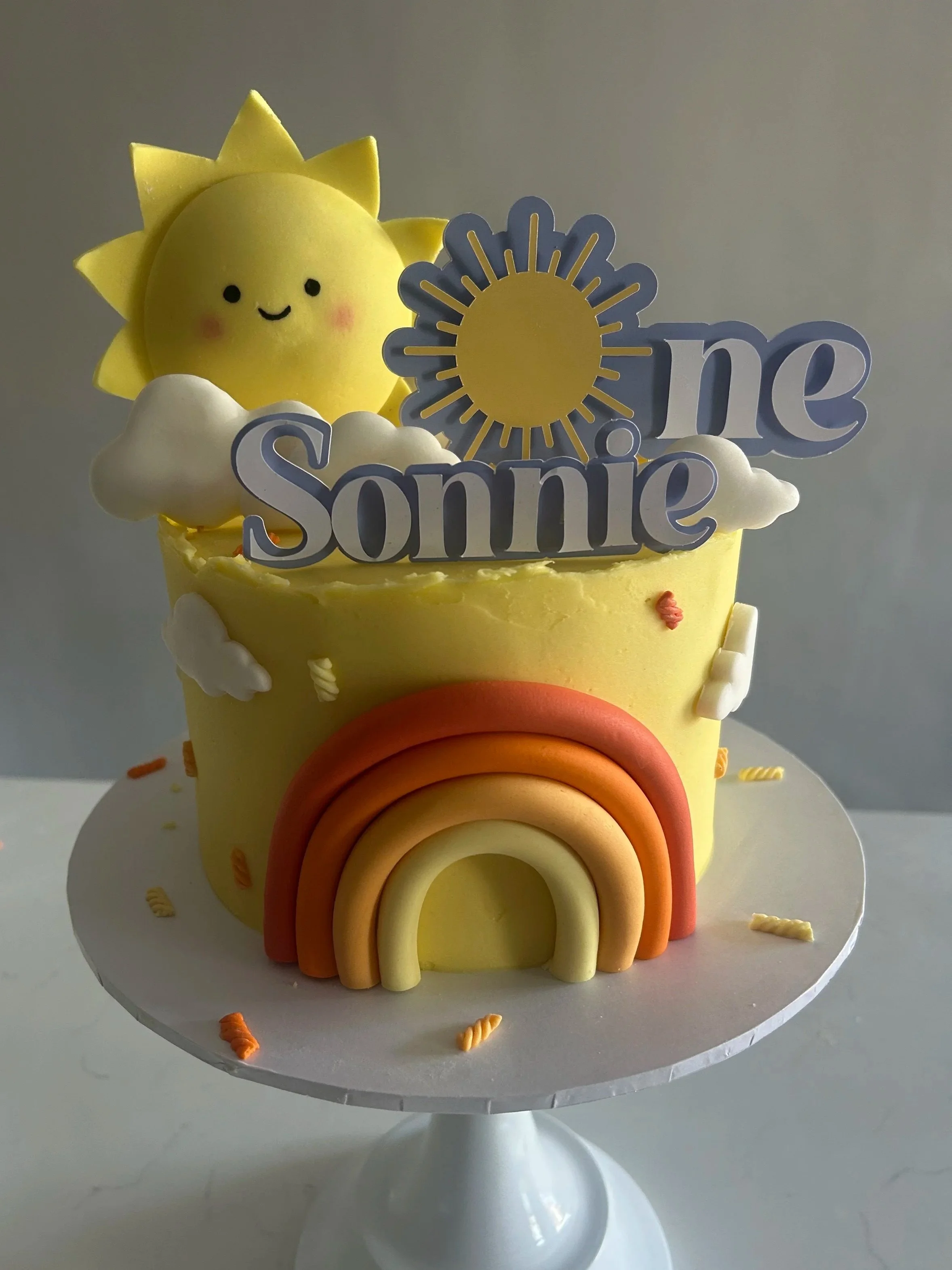 A yellow cake decorated with a smiling face sun topper, white cloud accents, and rainbow-colored fondant arches on a white cake stand, with a 'You Are My Sunshine' theme.