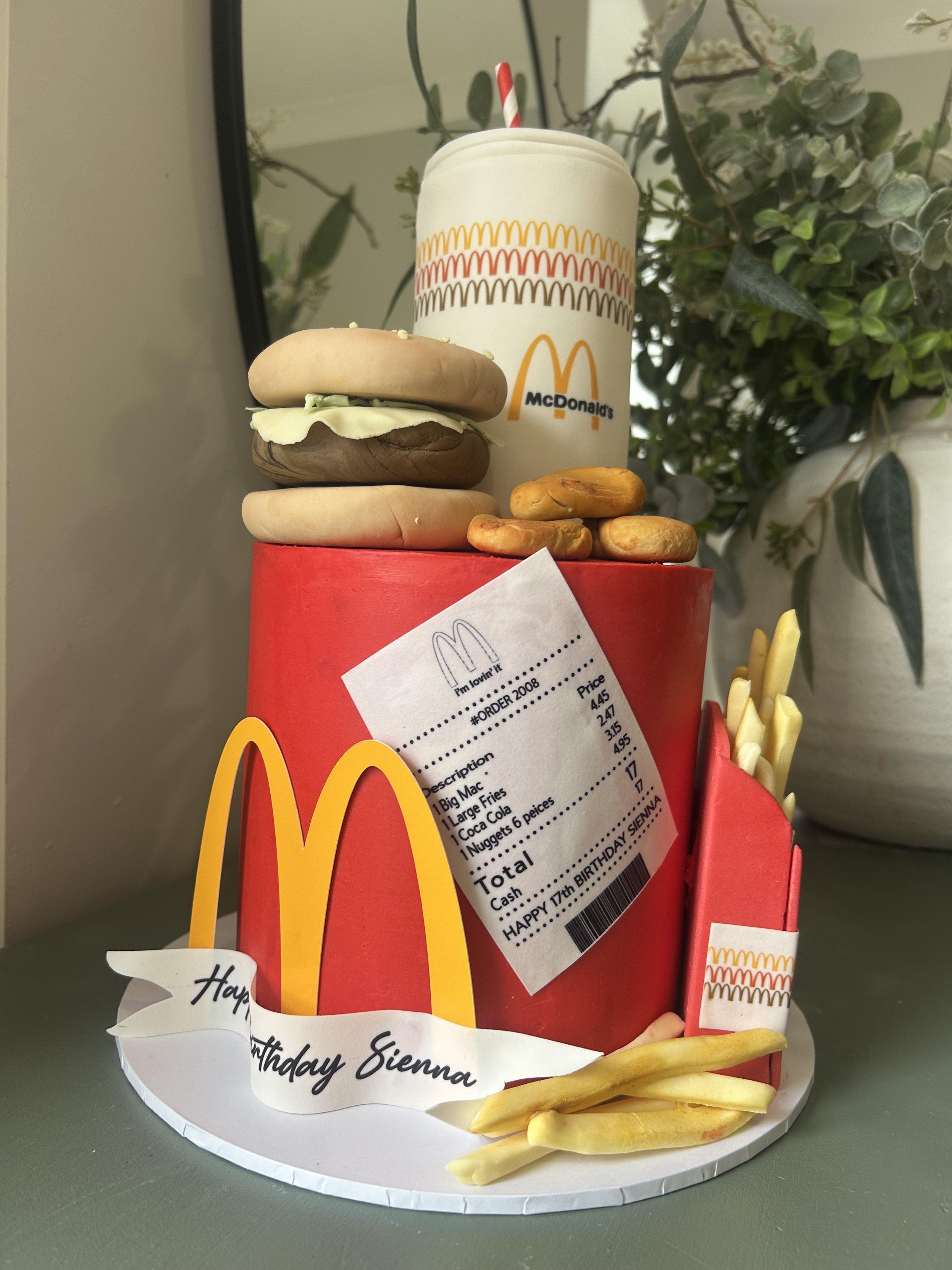 A McDonald's meal with a small fries box, a large soda cup, and three small sandwiches with buns, cheese, lettuce, and meat, placed inside a red container with a birthday note reading 'Happy Birthday Sienna' and a receipt attached.