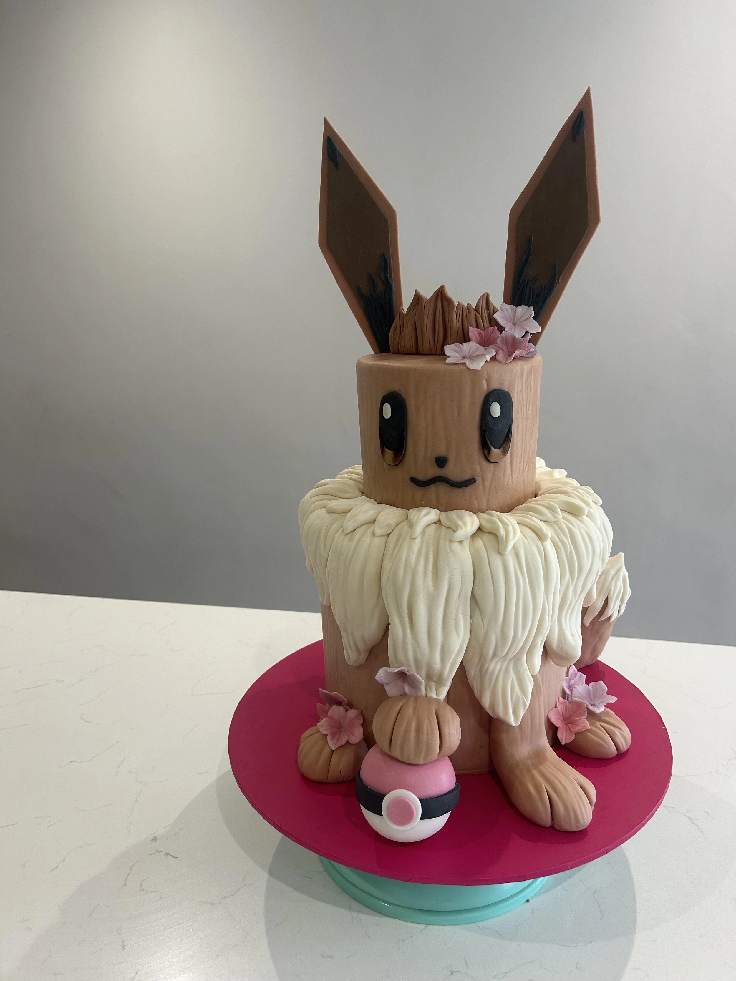 Cake decorated as Eevee from Pokémon, featuring large ears, big eyes, a small nose, and a cute mouth, with pink flowers and a Pokéball on a pink cake board.