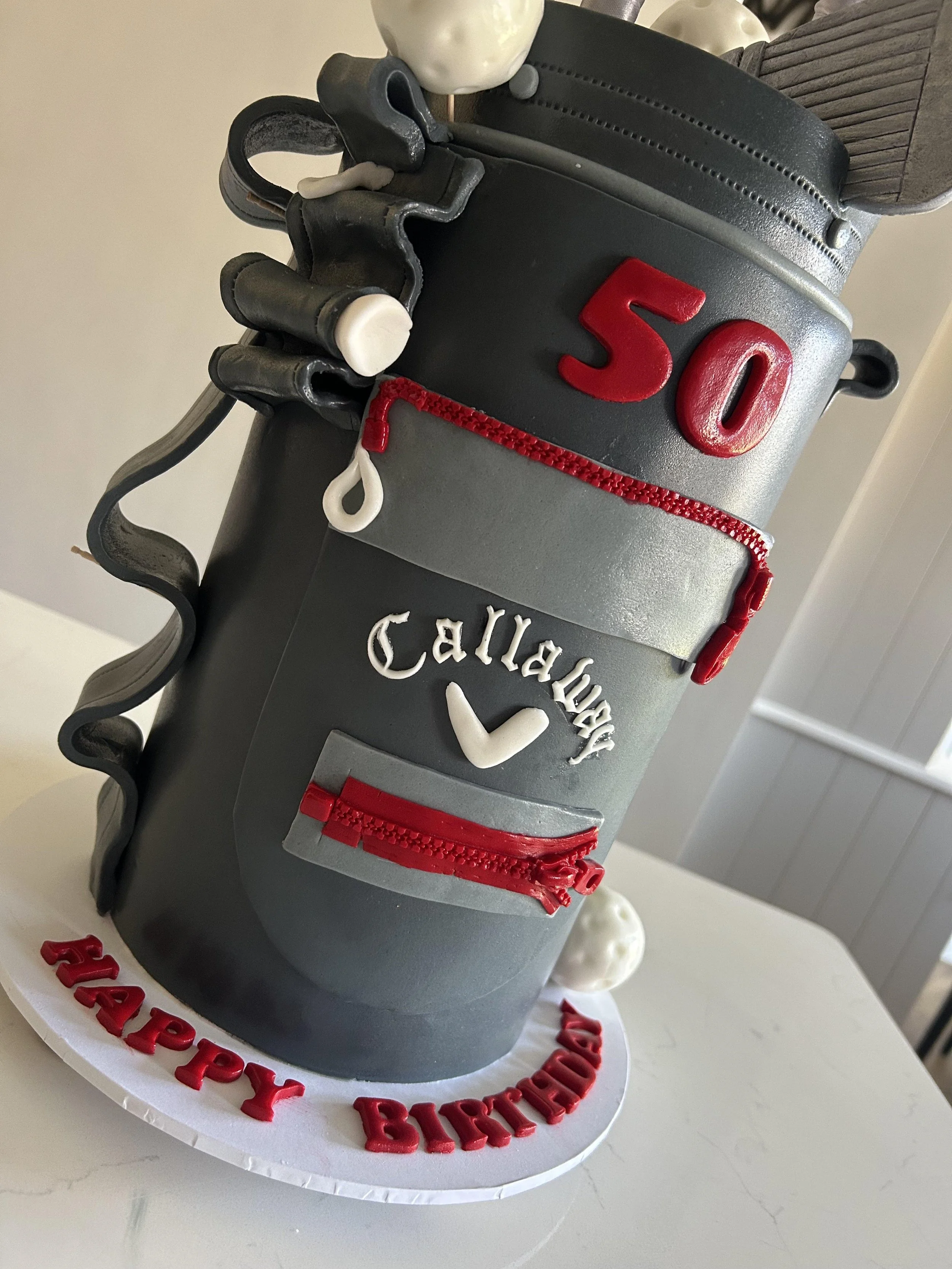 A cake decorated to look like a golf bag with clubs, a golf ball, and red details, with a birthday message in red lettering around the base.
