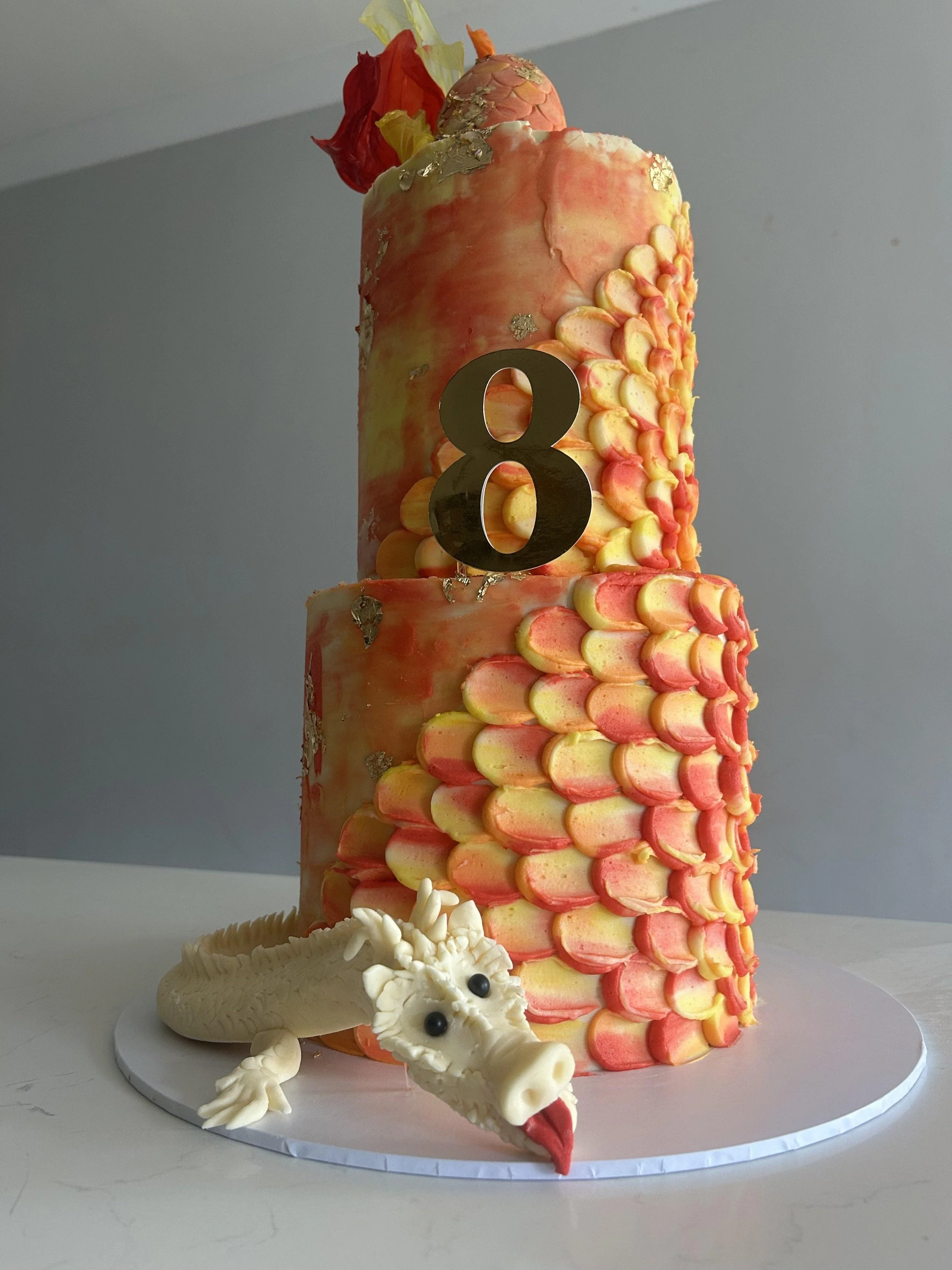 A multicolored cake decorated with a dragon figurine and a gold number 8 topper, with a textured base and bright, colorful icing patterns.