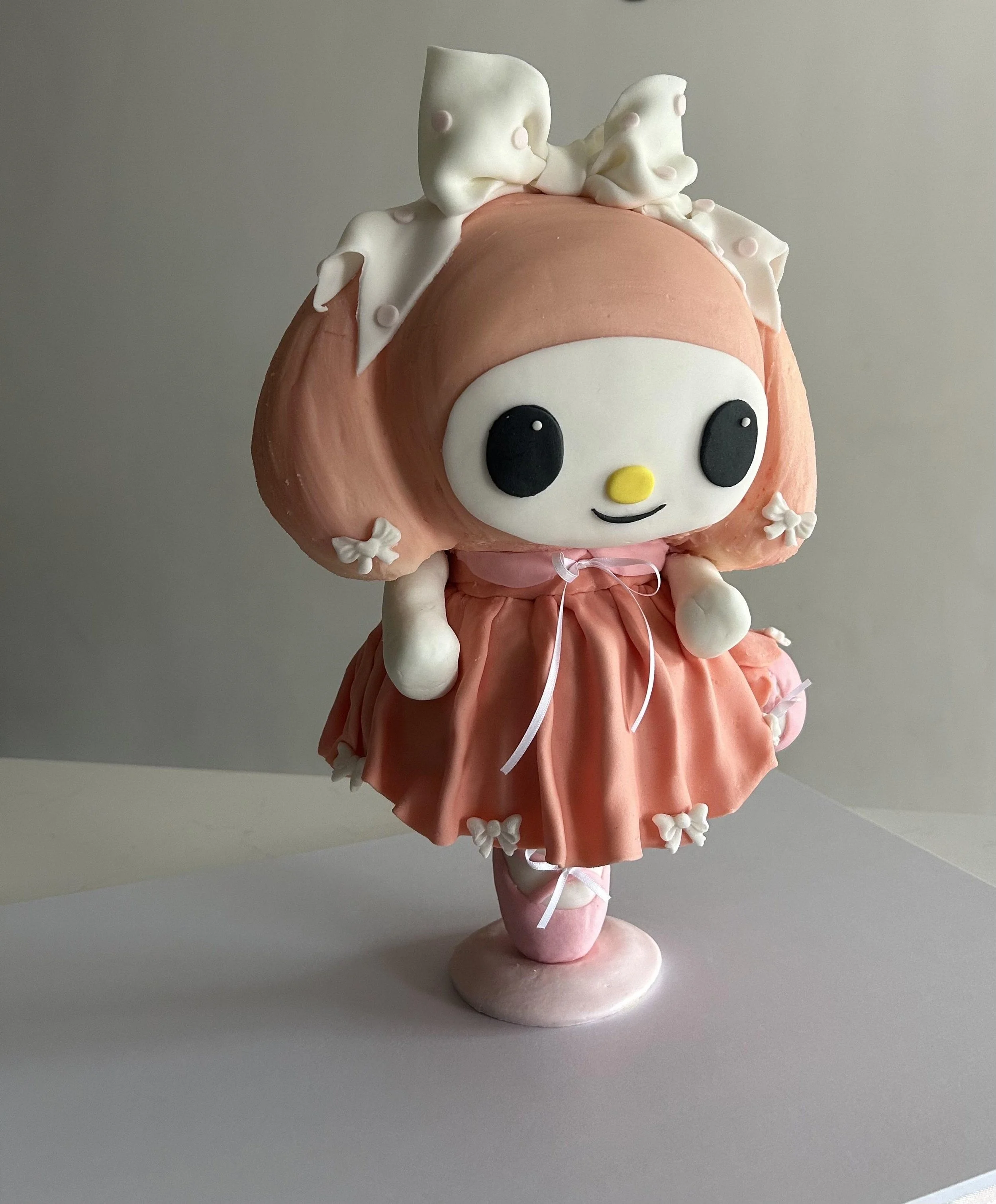 A Hello Kitty themed cake with a large round face, black eyes, yellow nose, small mouth, and a pink dress. The cake has white flower decorations and a plush Hello Kitty doll sitting on top, wearing a pink dress with bows and lace details.