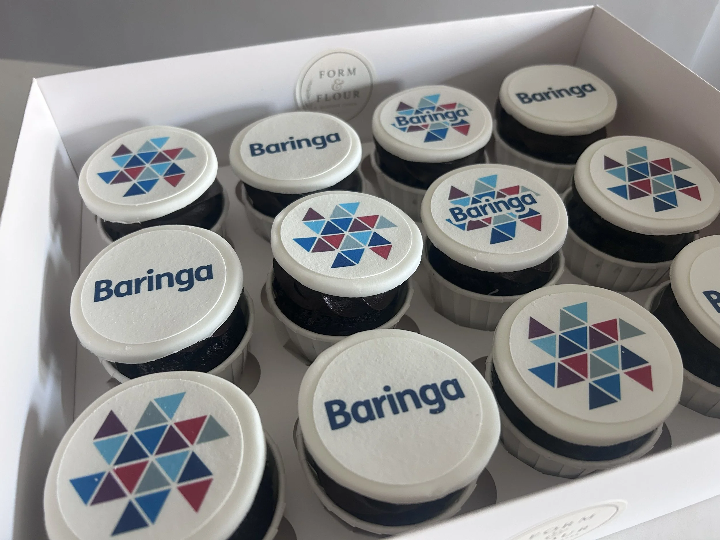 A white box containing several cupcakes, each with a white top and a logo with the word "Baringa" and a colorful geometric design.
