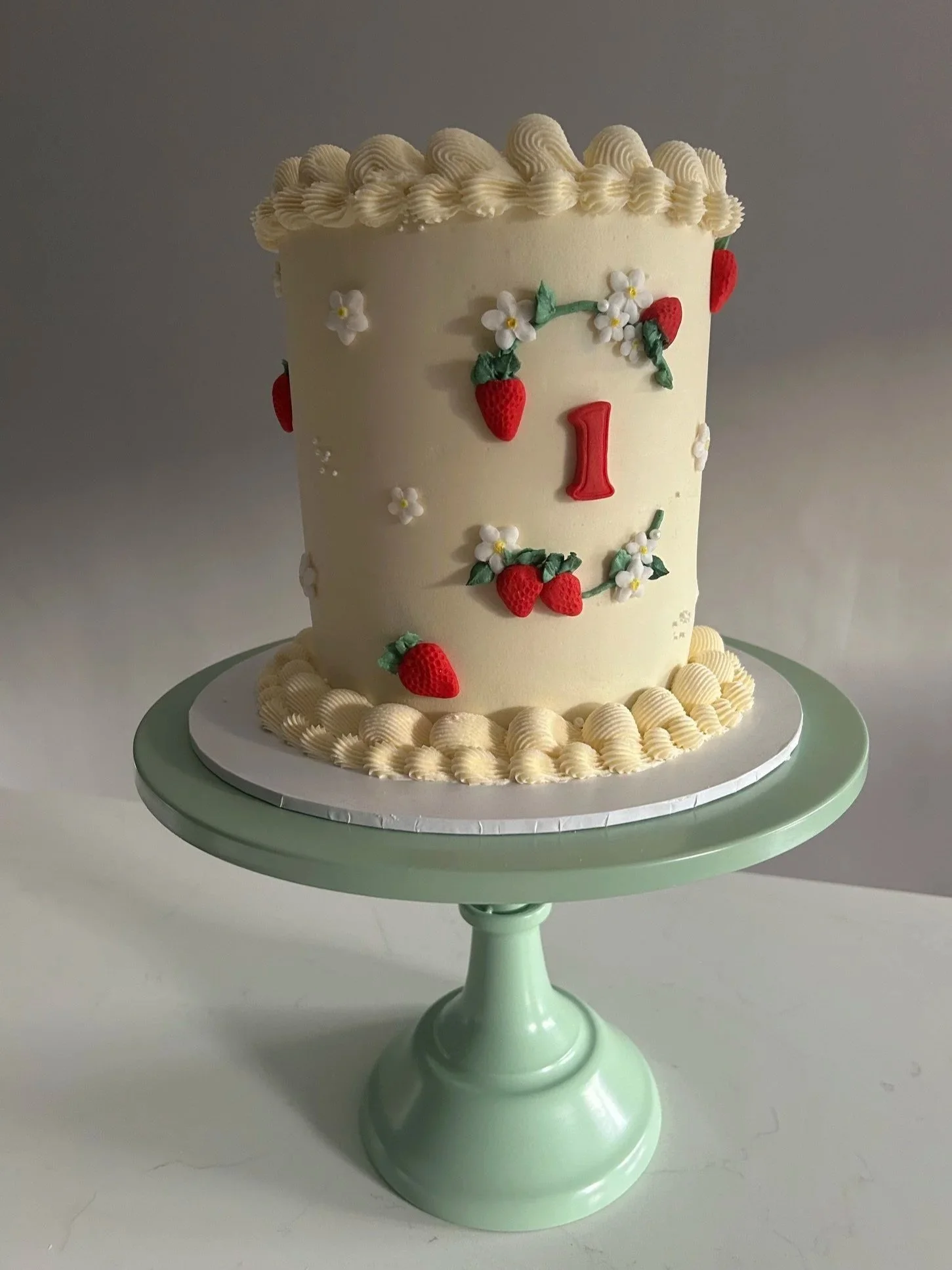 A tall, white birthday cake decorated with strawberries and white flowers, topped with piped white icing, on a light green cake stand.