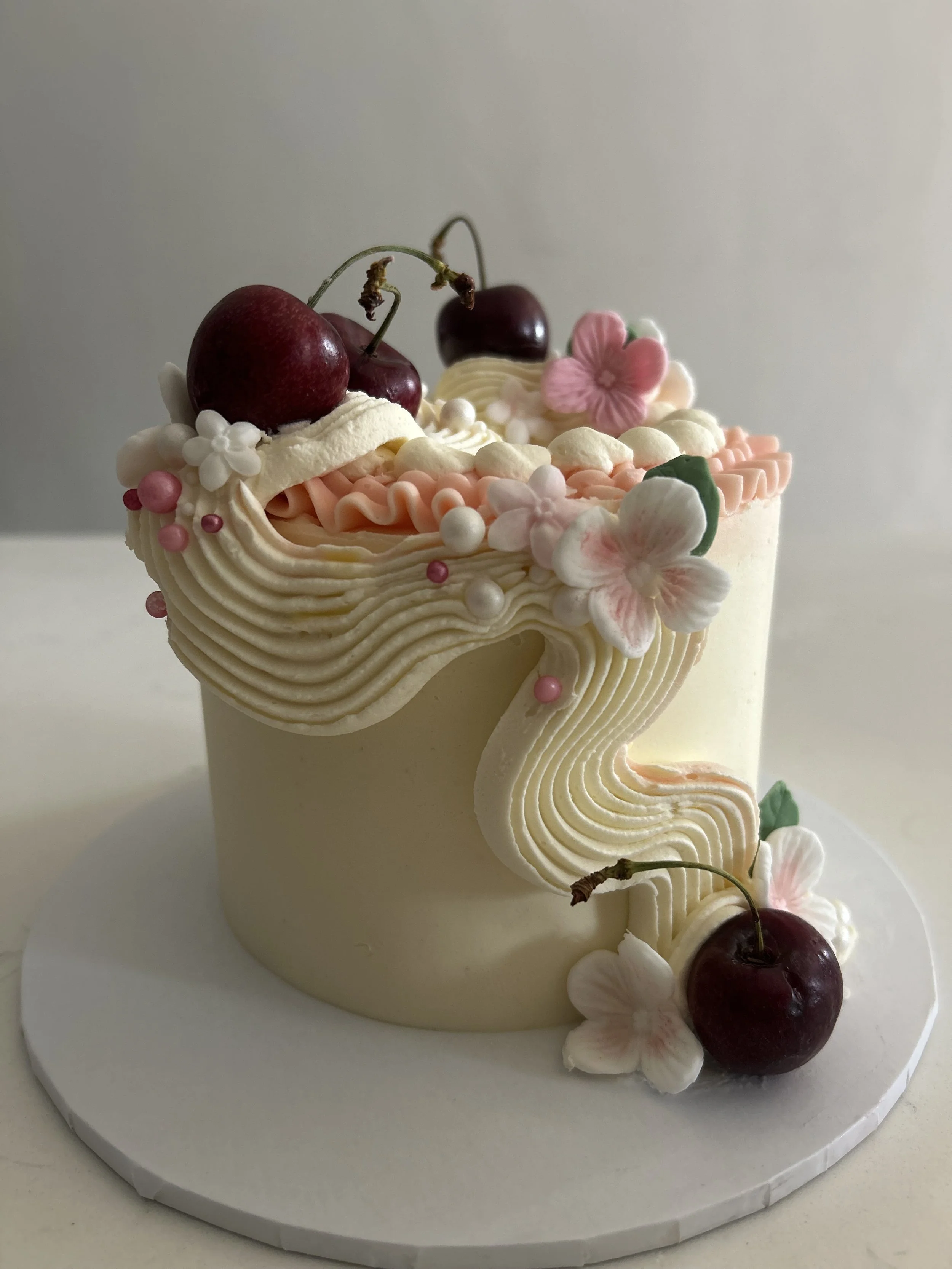 A decorative cake with cherries, pink and white flowers, and white and pink icing, with intricate piping and floral embellishments, on a white round base.
