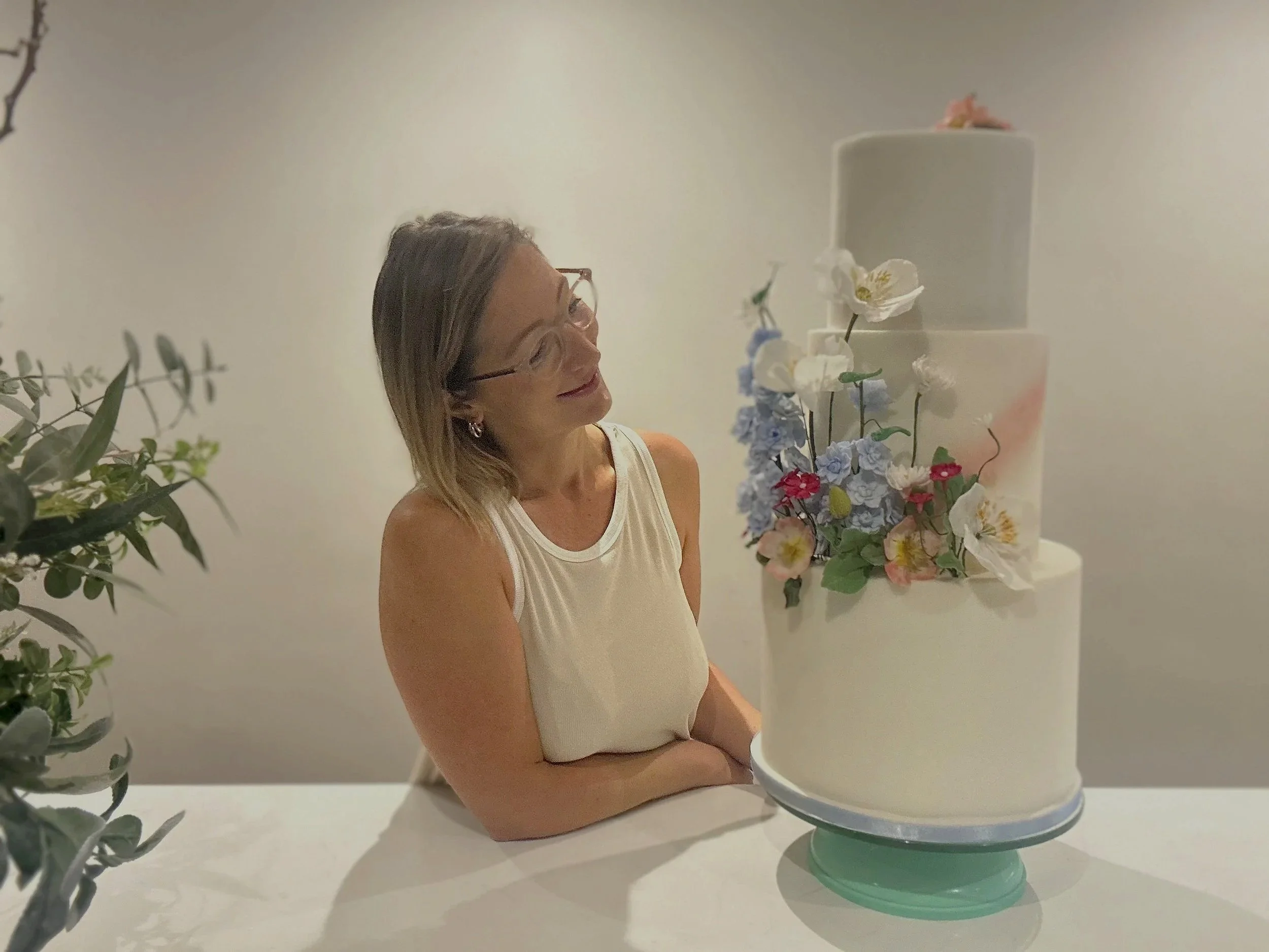 Rebecca; Form & Flour Cake Baker standing behind a multi-tiered white and pink cake on it, decorated with meadow-style flowers.