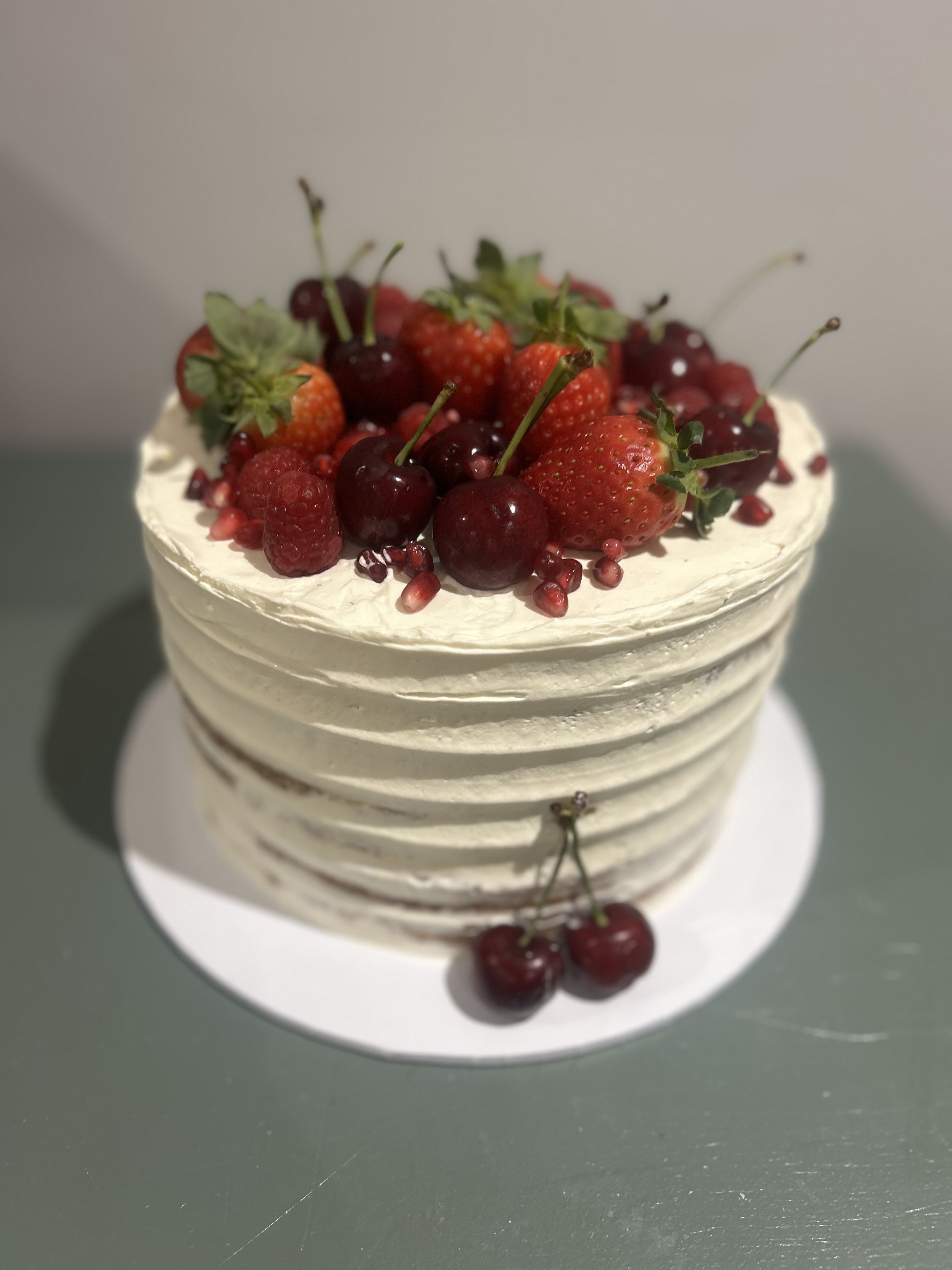 A layered cake topped with fresh strawberries, cherries, raspberries, and pomegranate seeds on a white cake board.