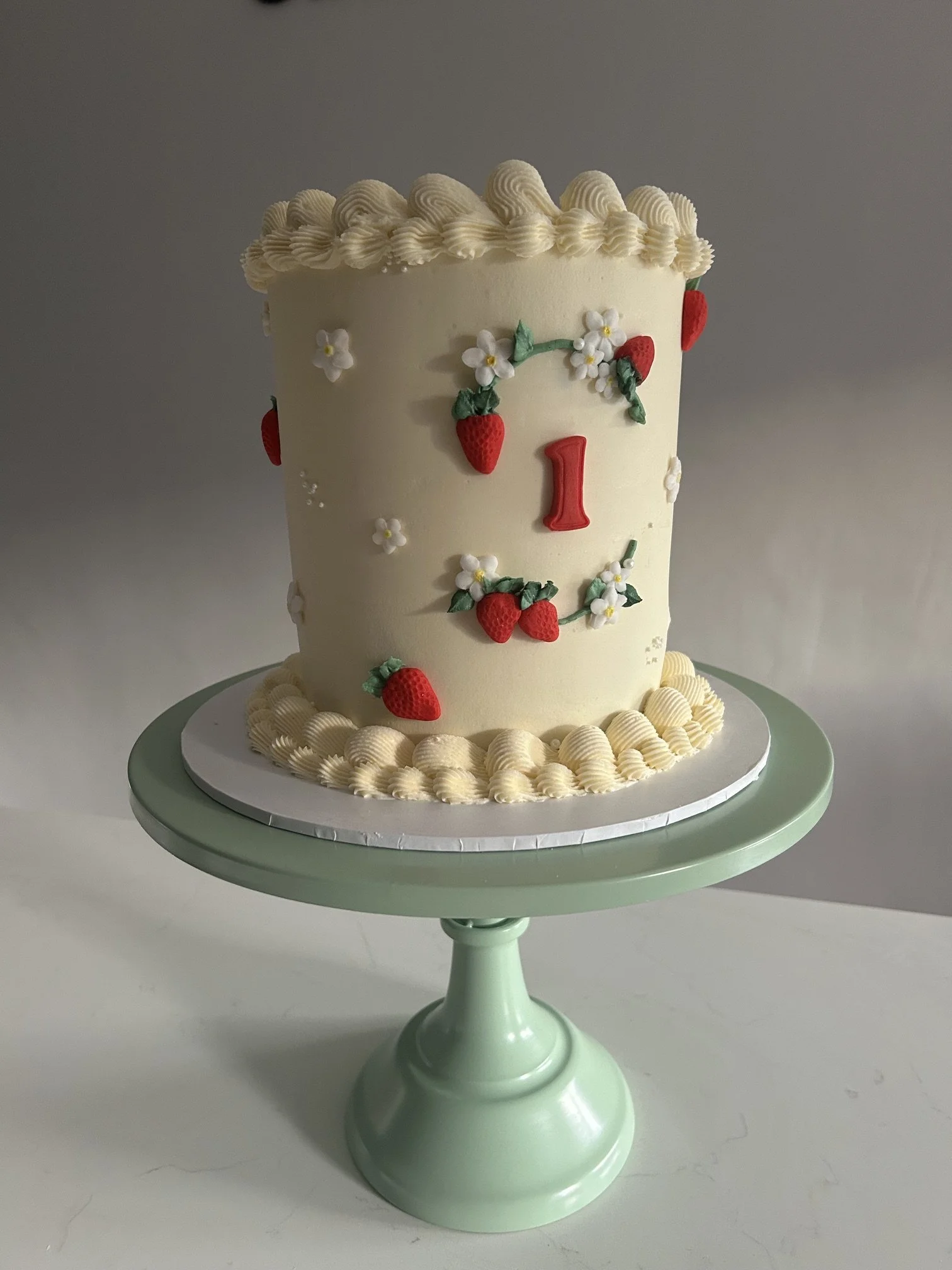 A tall birthday cake decorated with white icing and strawberries made of icing, featuring a red number 1 on the front, placed on a light green cake stand.