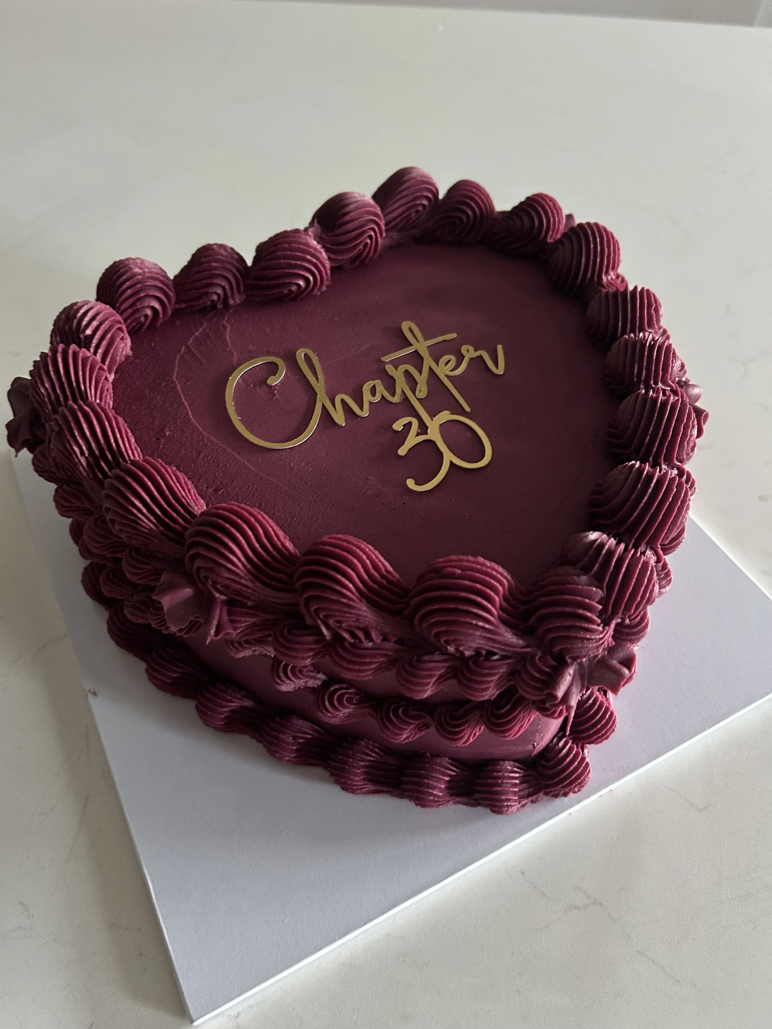 A round, pink anniversary cake with decorative pink frosting piping around the edge, featuring a gold 'Chapter 30' cake topper on top.