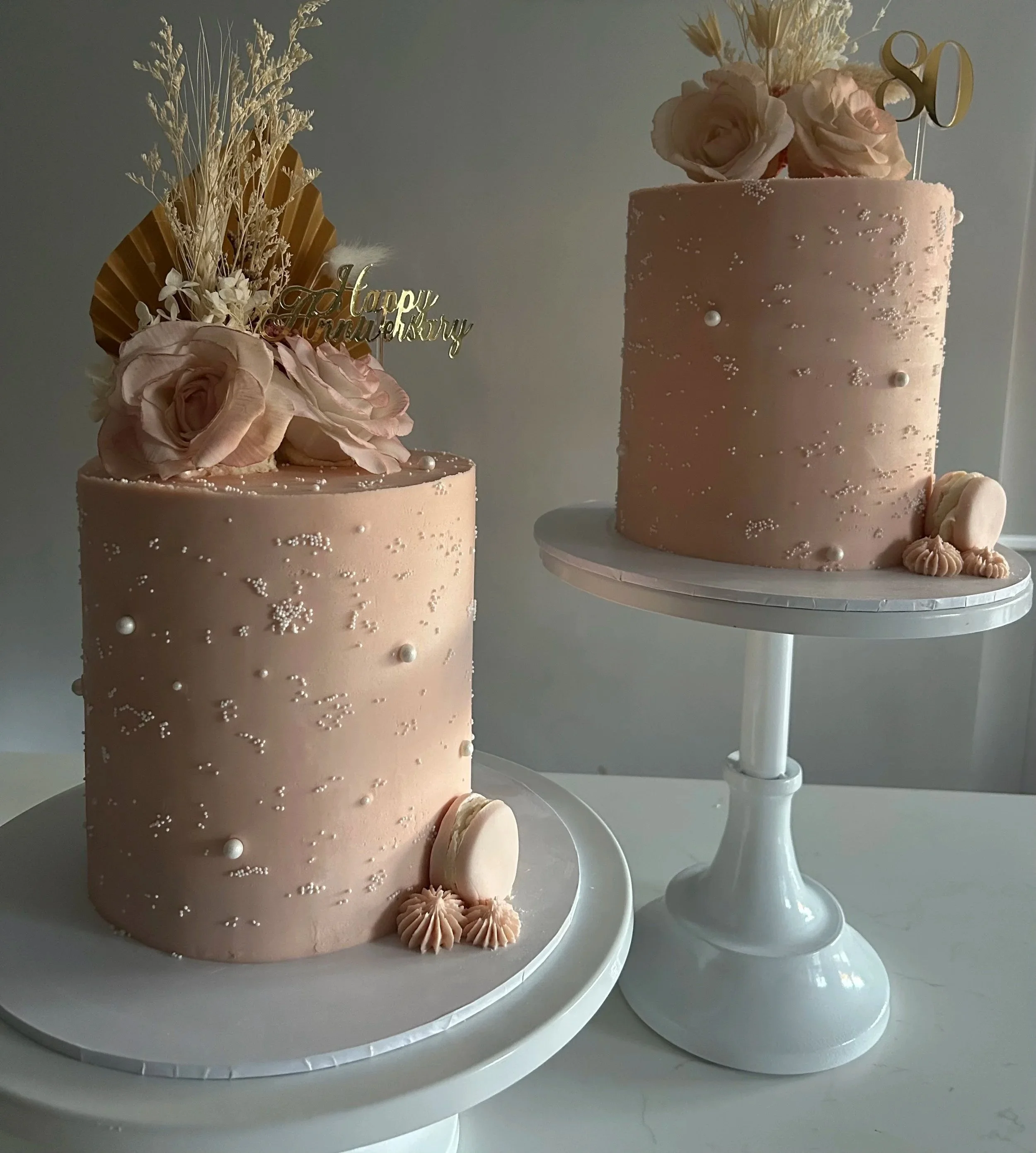 Two pink birthday cakes decorated with flowers, pearls, and sprinkles, each on a white cake stand, with a gold '50' cake topper and a gold 'Happy Birthday' sign.