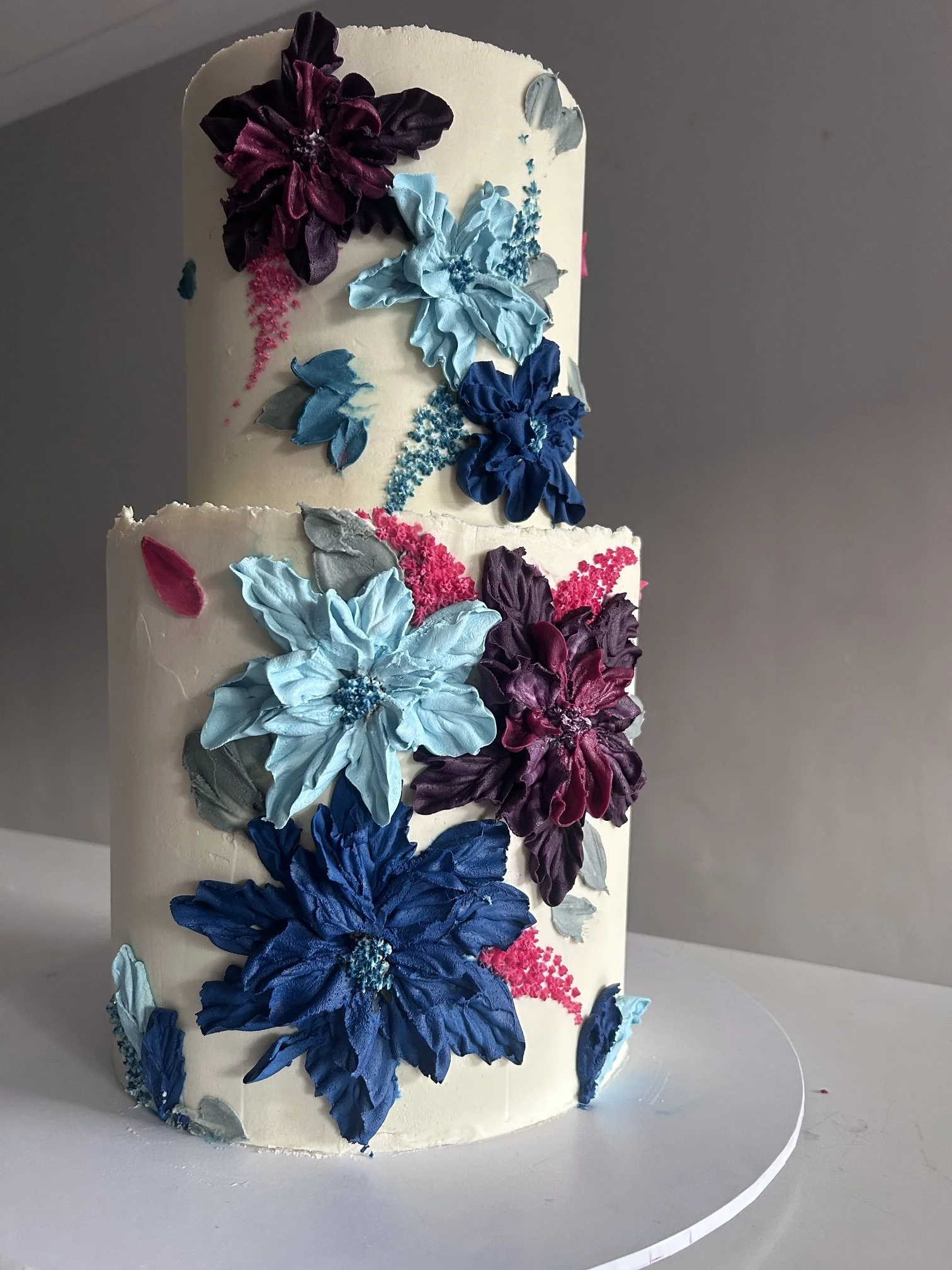 A white birthday cake decorated with large, colorful sugar flowers in shades of blue, purple, gray, and pink, arranged in vertical lines around the cake.