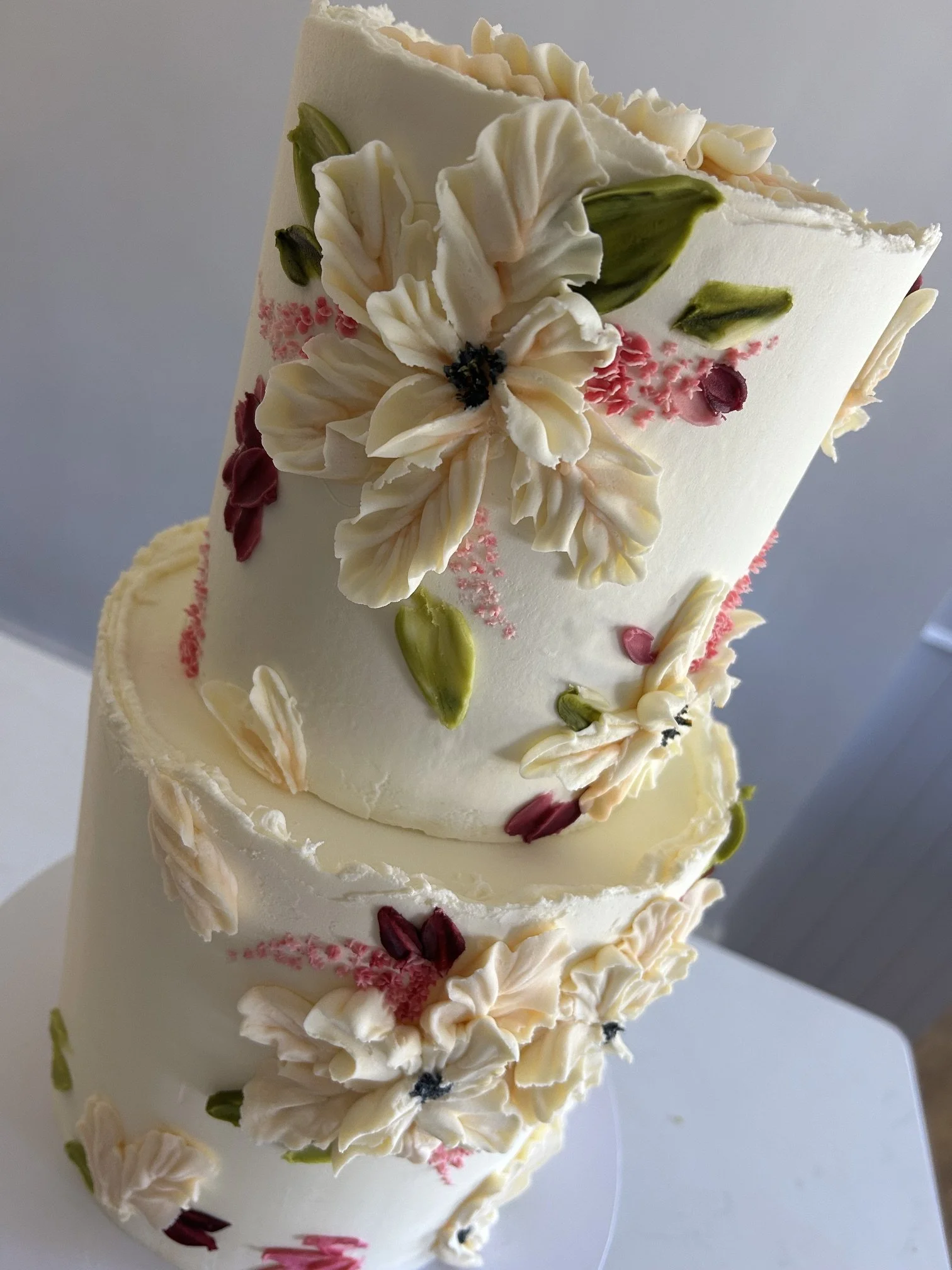 Close-up of a tall, elegant white cake decorated with white, pink, and green floral icing, featuring detailed flower designs and piped borders.
