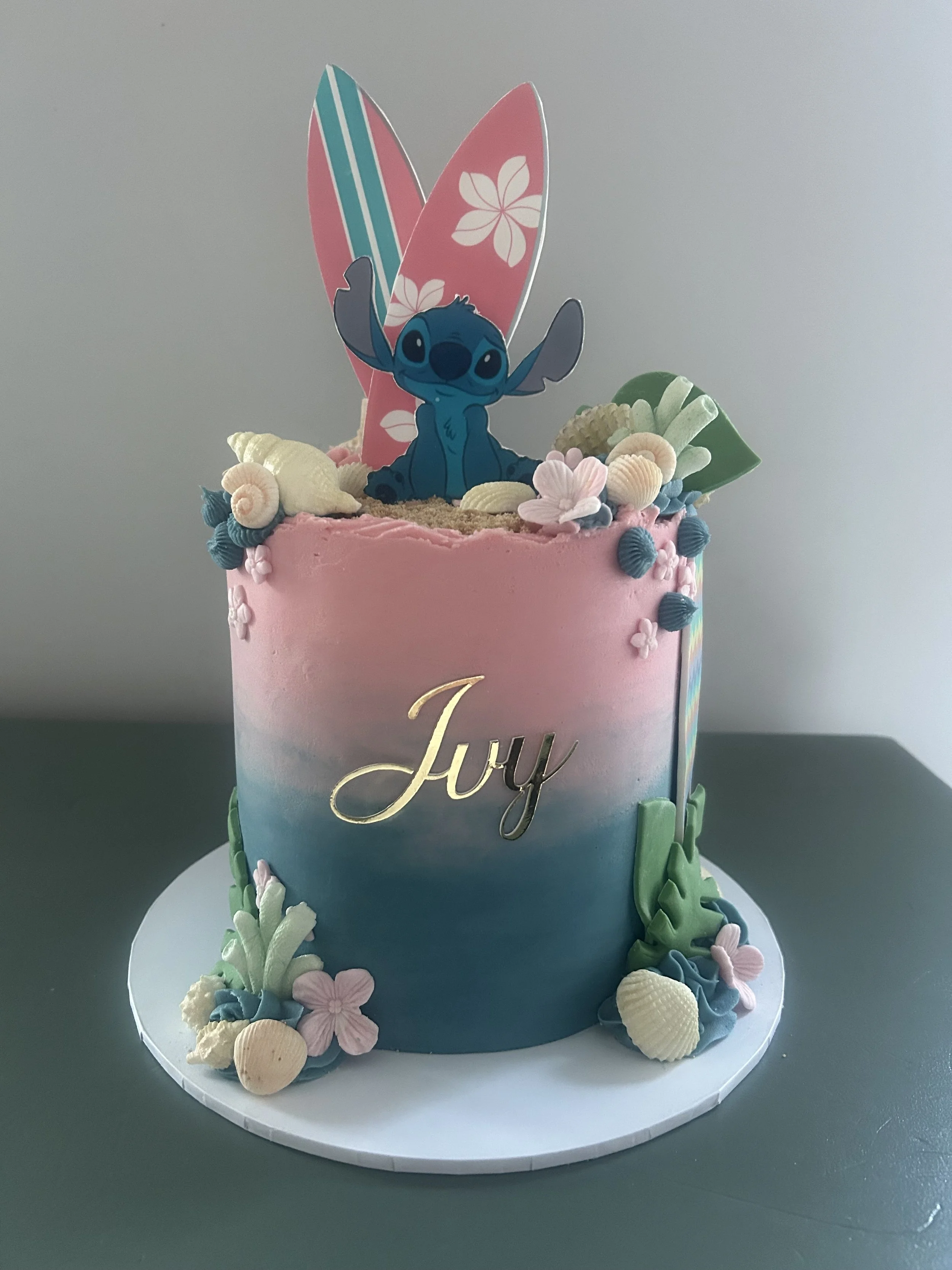 A colorful birthday cake with an edible figure of Stitch from the movie Lilo & Stitch on top. The cake is decorated with seashell-shaped candies, flowers, and pastel-colored frosting, with parts of the cake showing an ombré effect from pink to blue.