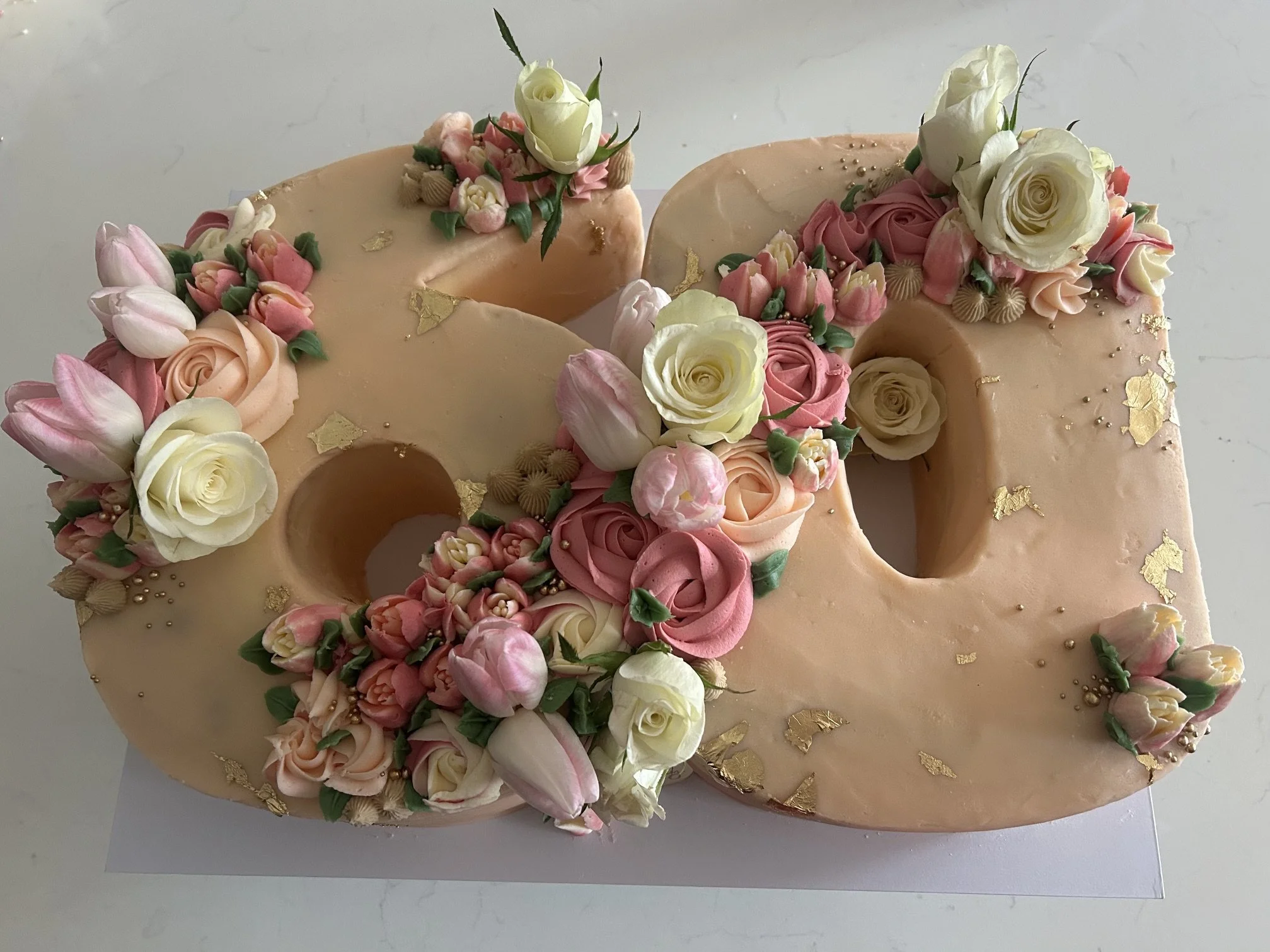 Close-up of a blush pink cake decorated with light pink, cream, and white roses, tulips, and small flowers, adorned with gold leaf and tiny edible pearls.