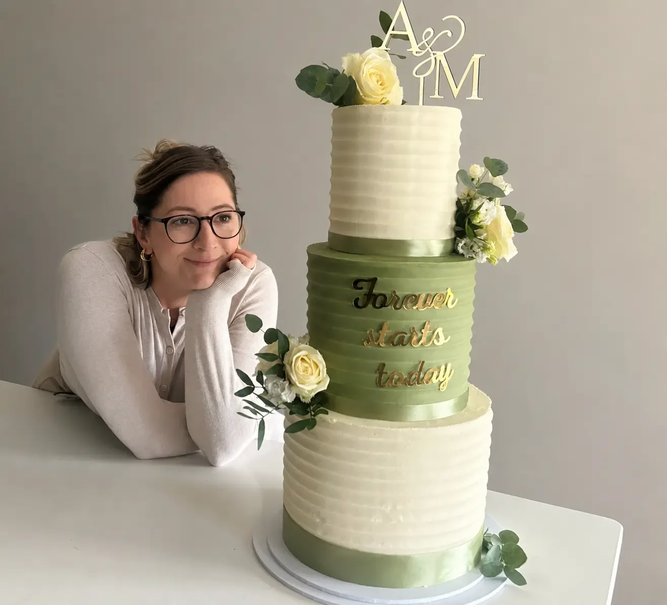 multi-tiered wedding cake decorated with white and green flowers, with a topper of intertwined letters and the words "forever starts today" on the middle tier.