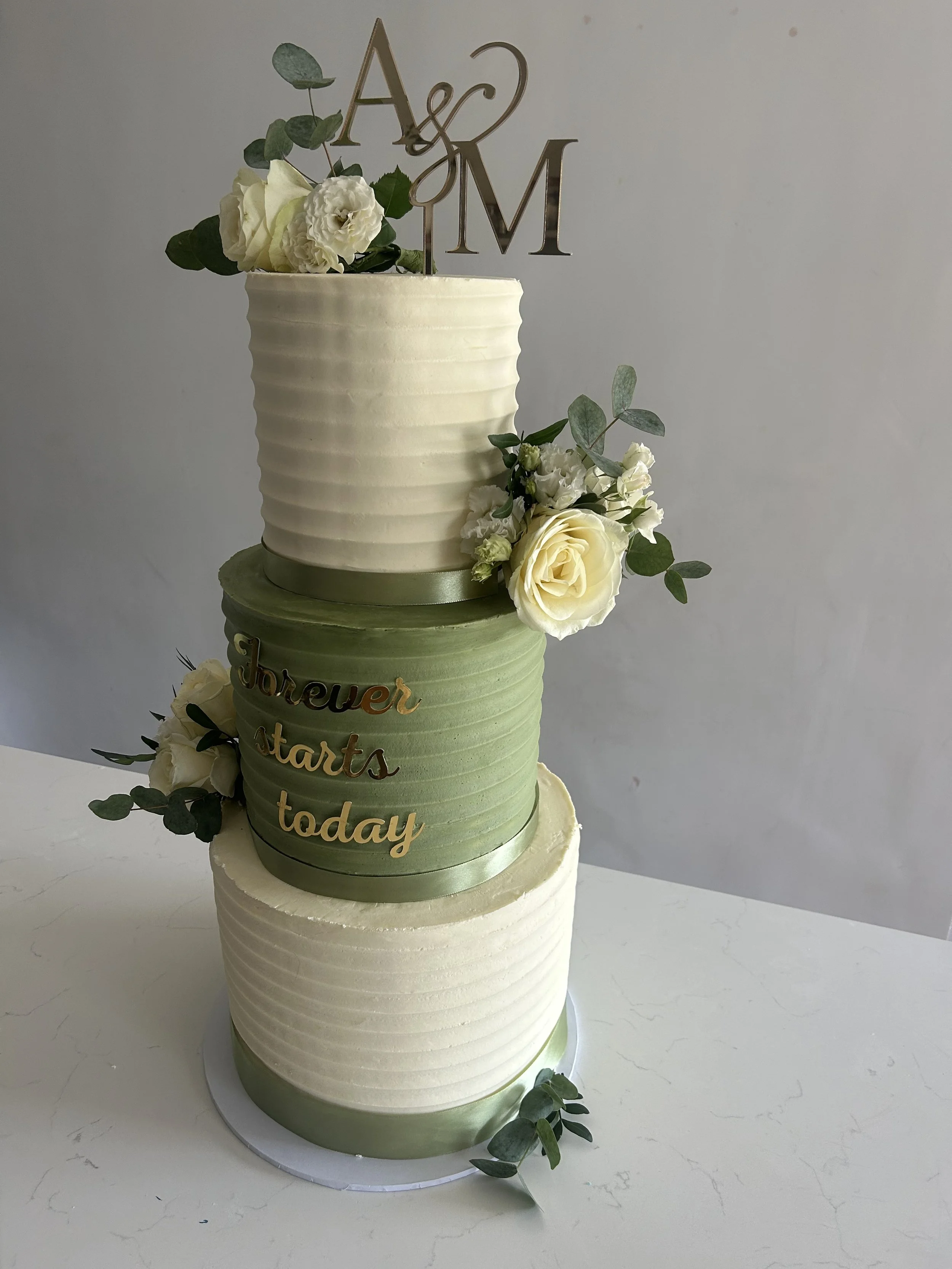 A three-tier wedding cake decorated with white flowers, green leaves, and a silver topper with the initials 'A & M'. The cake has a light green ribbon at the bottom of each tier and a gold-colored inscription that reads 'Forever starts today' on the 